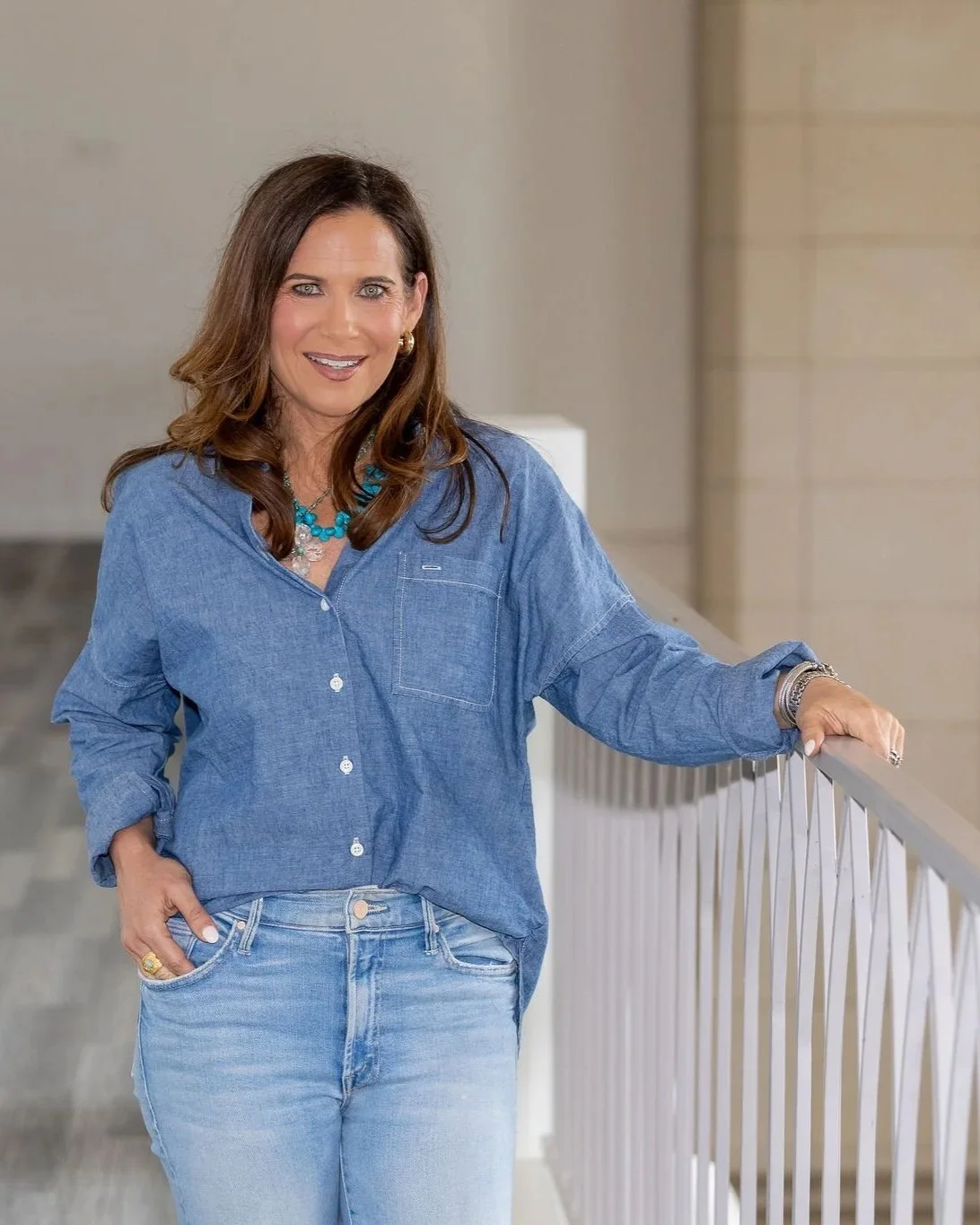 Woman with brown hair in a blue denim shirt and light blue jeans standing by a white railing indoors, smiling at the camera.