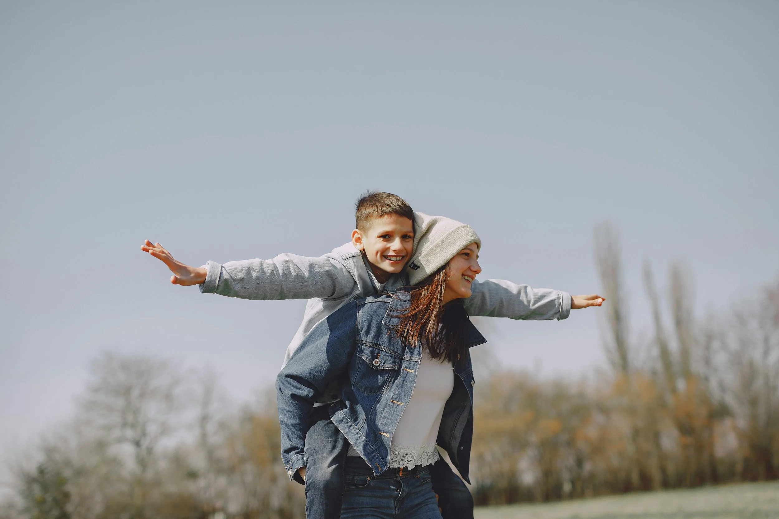 A woman carrying a young boy on her back outdoors, both smiling and extending their arms outward against a background of trees and a cloudy sky.