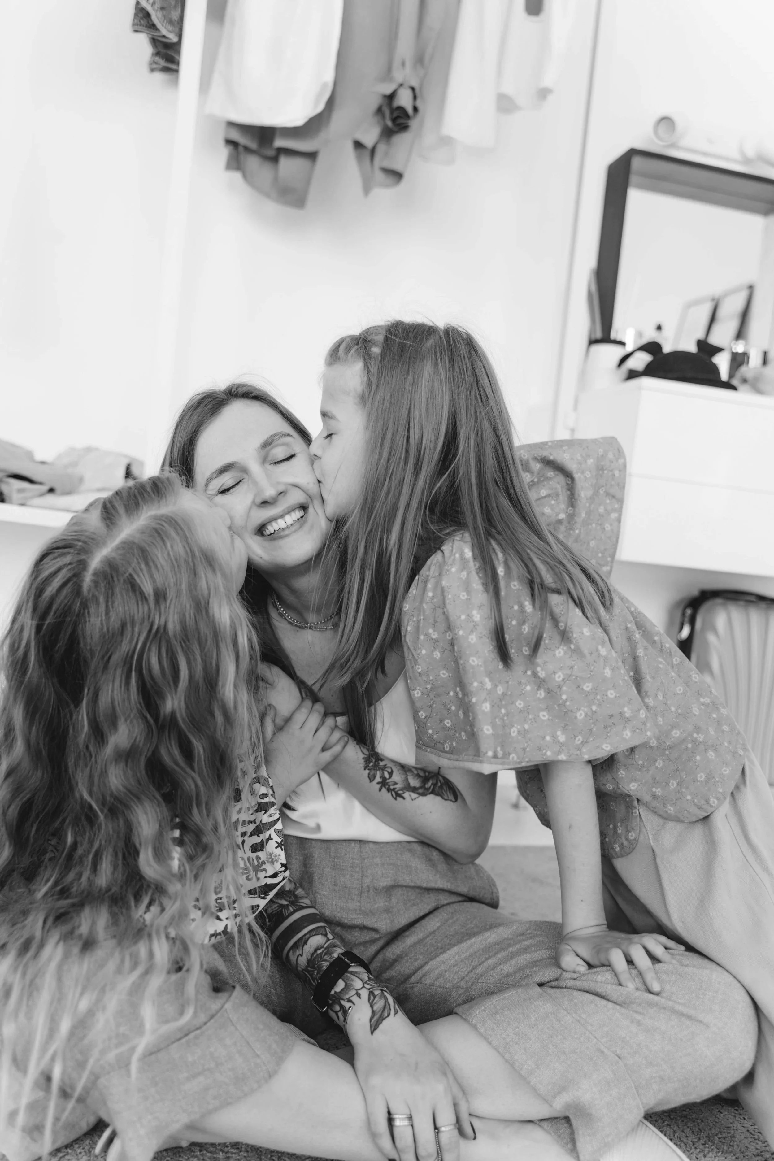A woman with long hair and tattoos on her arm smiles happily as she is kissed on the cheek by a young girl. Another younger girl with curly hair is also kissing her. The woman is seated on the floor with her legs crossed, and they are in a room with a mirror, clothes hanging, and some household items in the background.