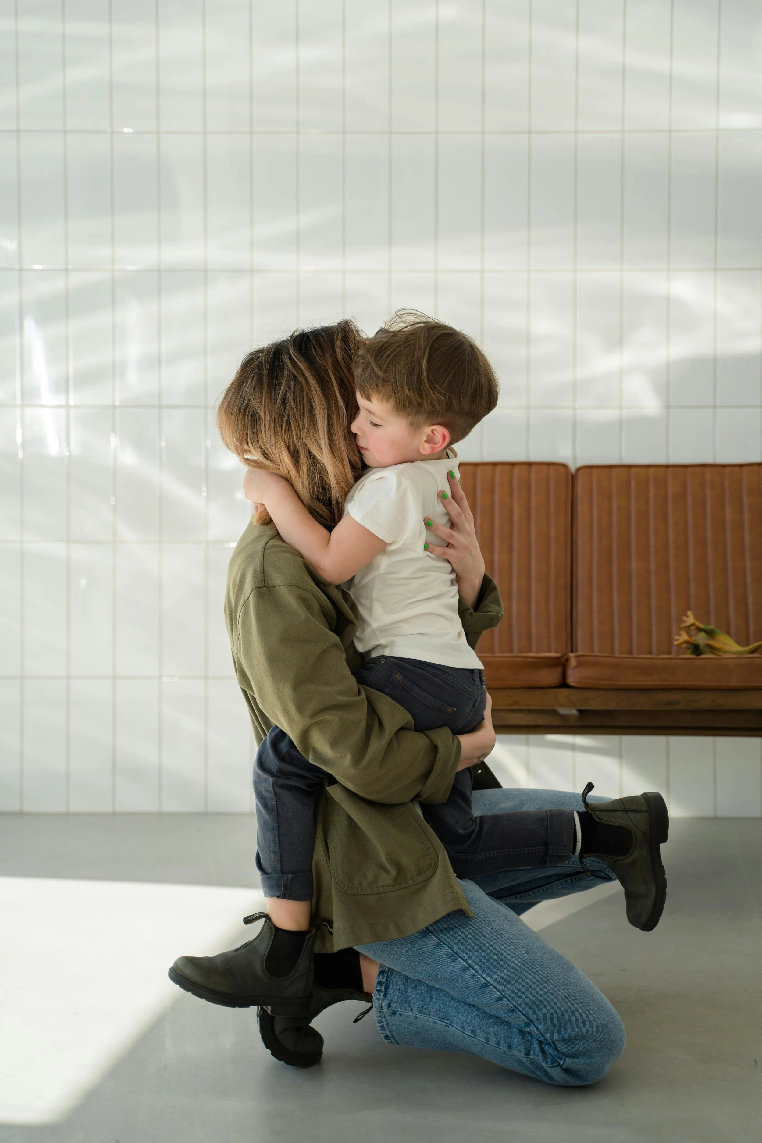 A woman is kneeling on the floor with her eyes closed, hugging a young boy with brown hair, who is sitting on her lap and hugging her back. They are in a room with white tiled walls and a wooden bench in the background.