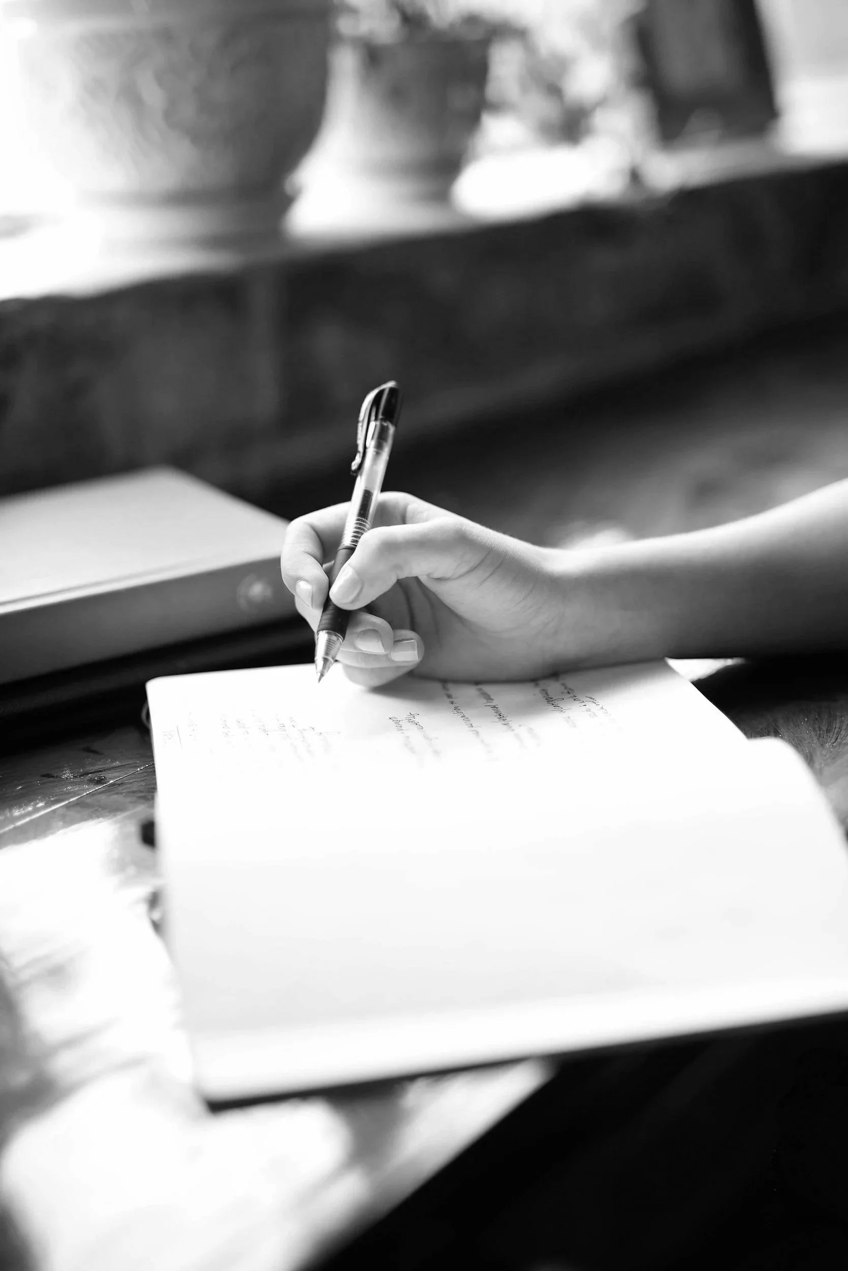 A person's hand is holding a pen and writing on a handwritten notebook. The background is blurred, showing a desk and some objects.