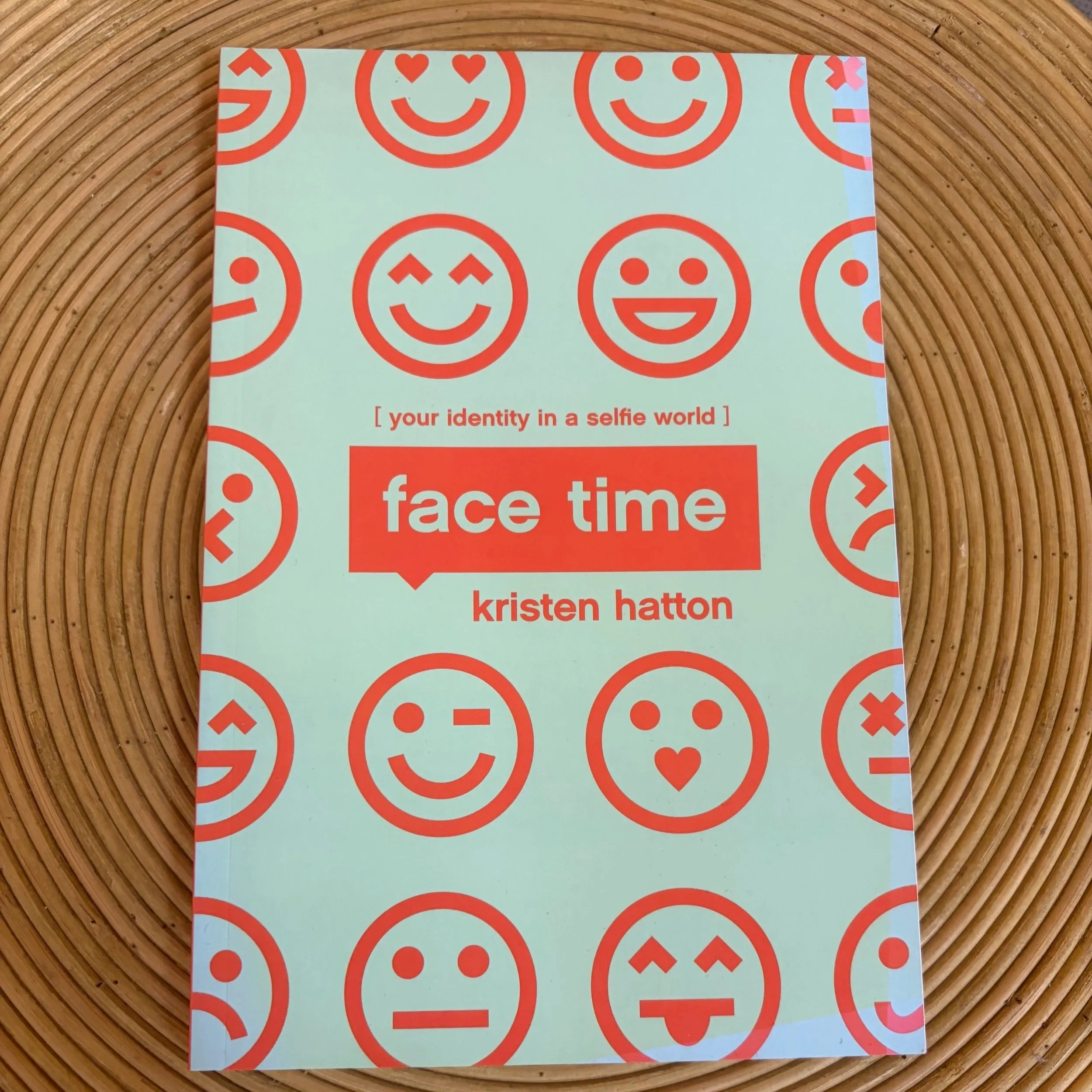 A book titled 'face time' by Kristen Hatton features a cover with multiple red smiley faces on a pale green background. The book is placed on a circular wooden surface.