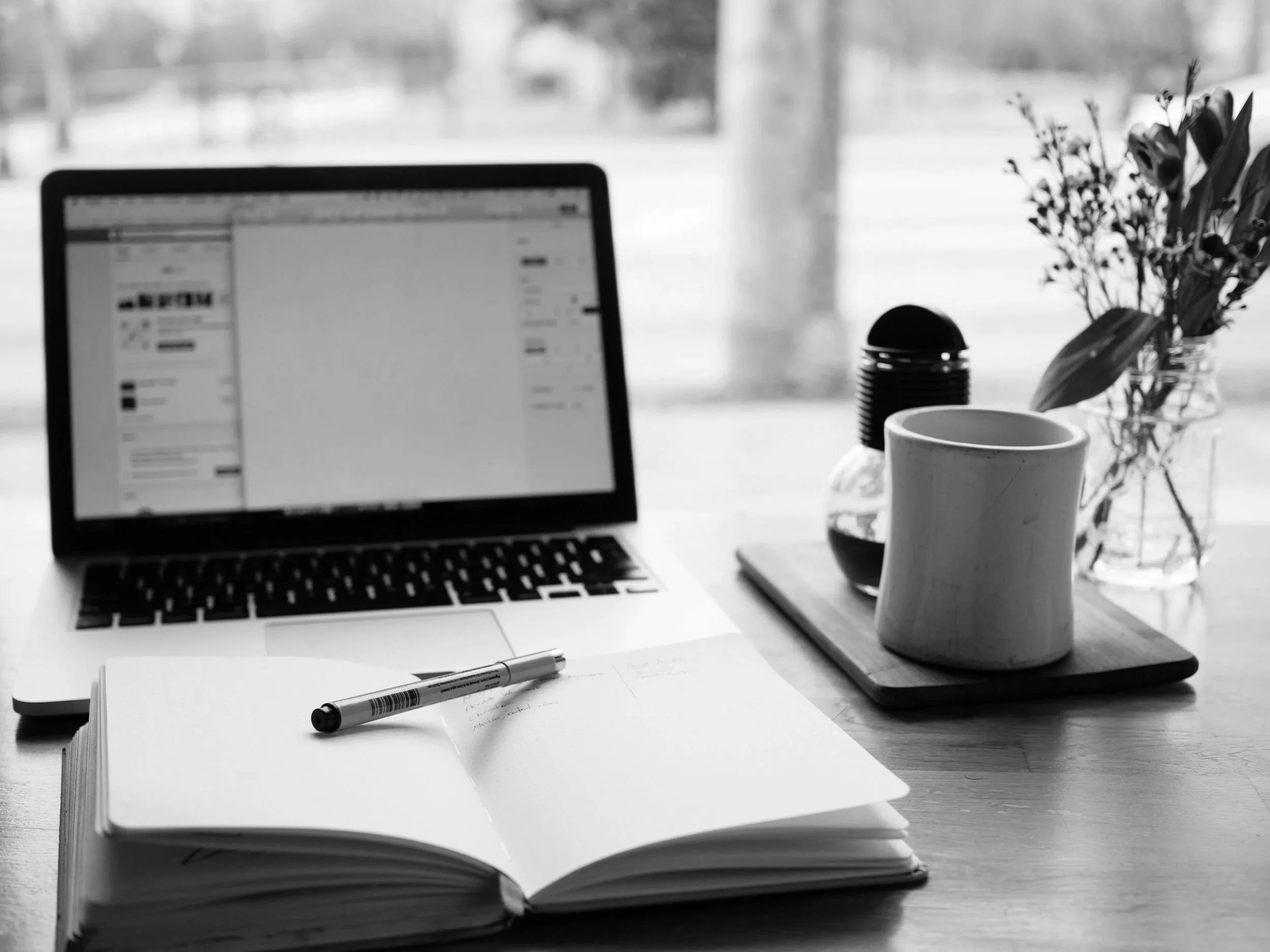 A workspace with a laptop displaying a social media page, a notebook with notes, a marker, a mug, a small jar of flowers, and a closed notebook on a wooden table, with a blurred background of a window and curtains.