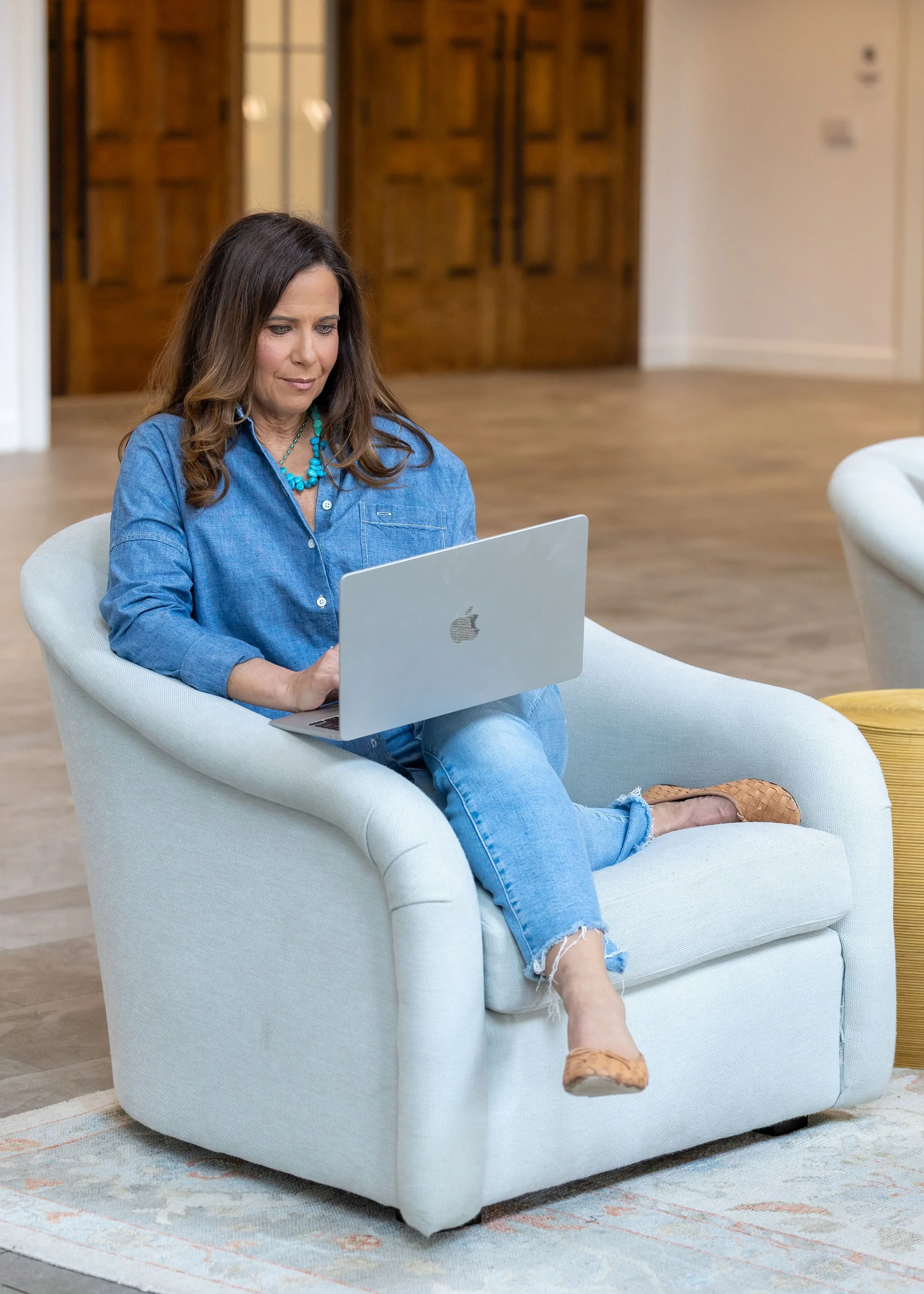 Woman sitting on a light-colored chair, working on a silver laptop in a room with wooden doors, a beige wall, and a patterned rug.
