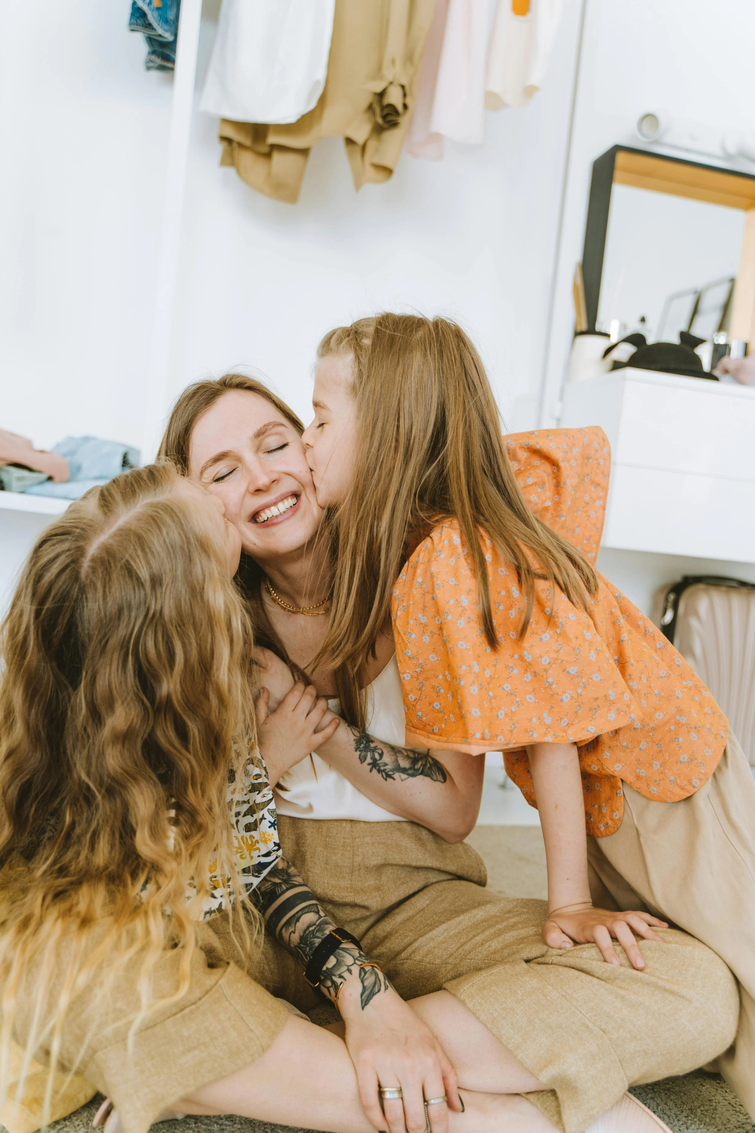 A woman with long, light brown hair smiling with eyes closed, surrounded by children, and a girl giving her a kiss on the cheek.