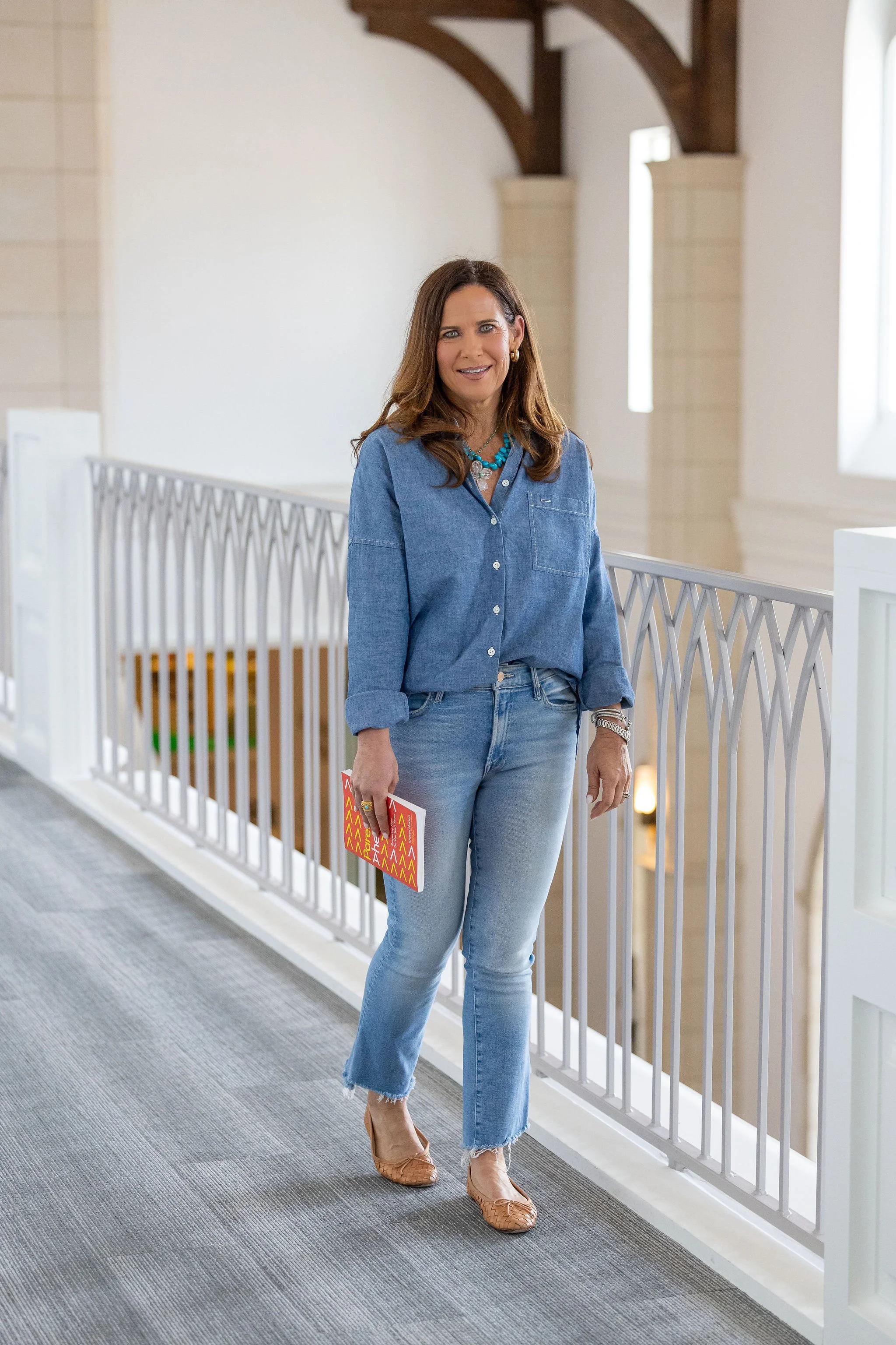 Woman standing in a bright, airy space, holding a brochure, wearing a denim shirt and jeans, with wooden beams and large windows in the background.