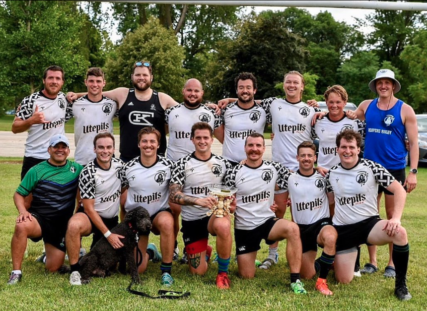 A group of young male rugby players in team uniforms posing outdoors with a coach and a dog, after a game or practice, on a grassy field with trees in the background.