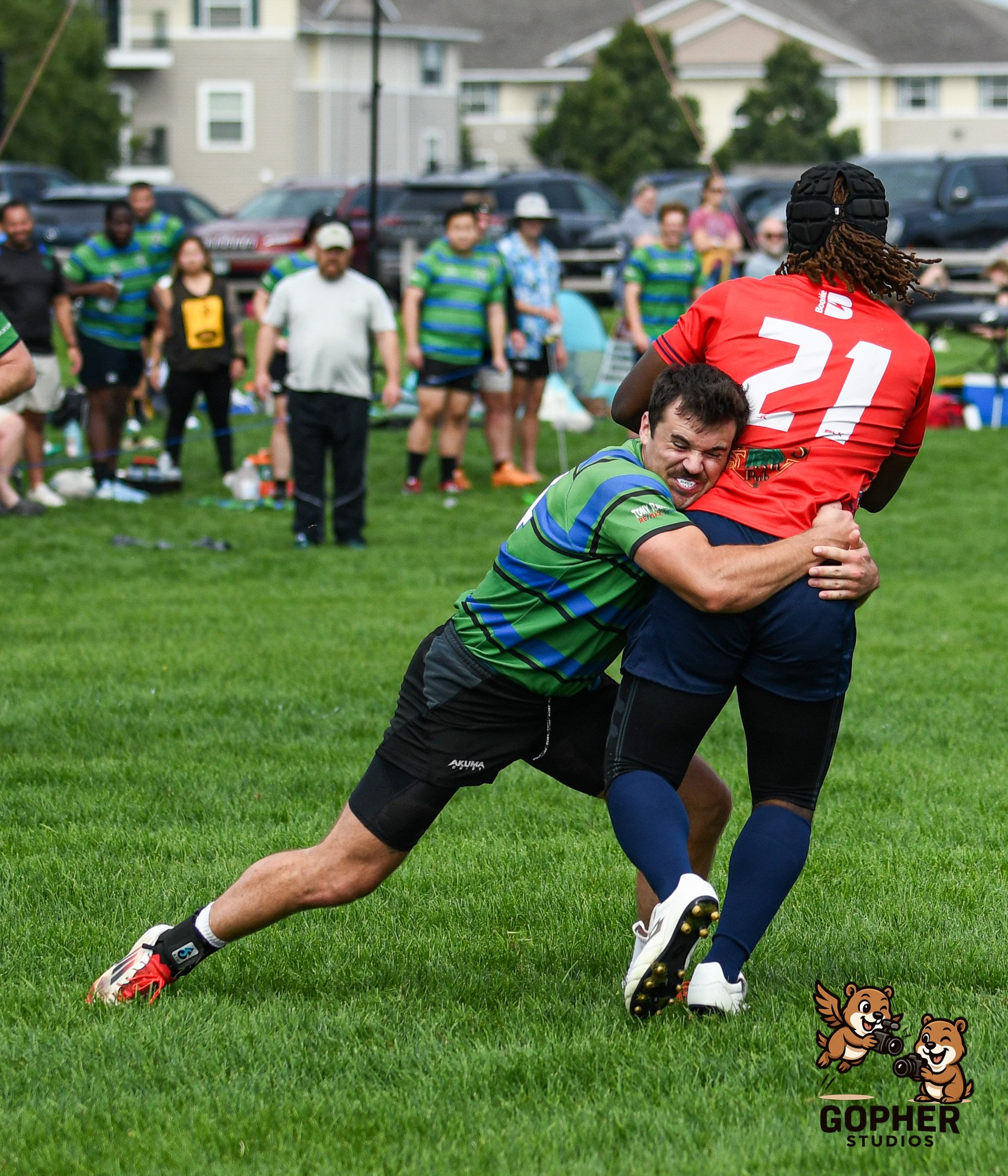 A rugby game with two players tackling each other on a grassy field, with spectators watching in the background. One player is wearing a red jersey with the number 21, and the other player is in a green and blue striped jersey. There is a Gopher Studios logo with cartoon gophers holding cameras in the lower right corner.