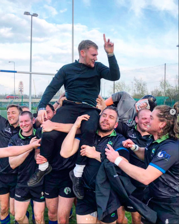 A group of rugby players celebrating on a field, lifting a man in black clothing in the air, who is raising one finger in victory.