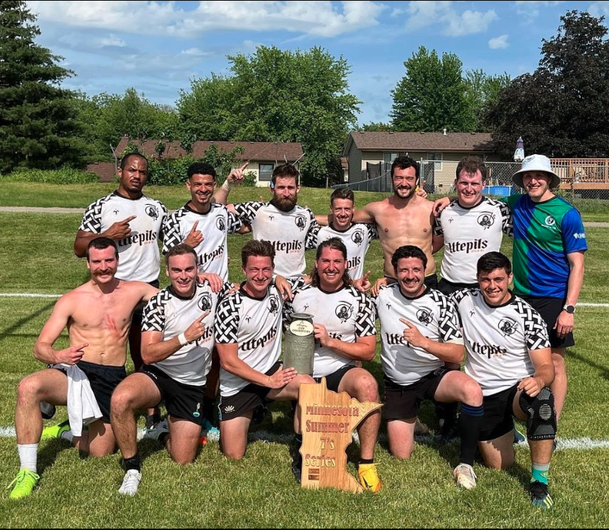A soccer team celebrating after winning a match, holding a trophy, on a grassy field with houses and trees in the background.