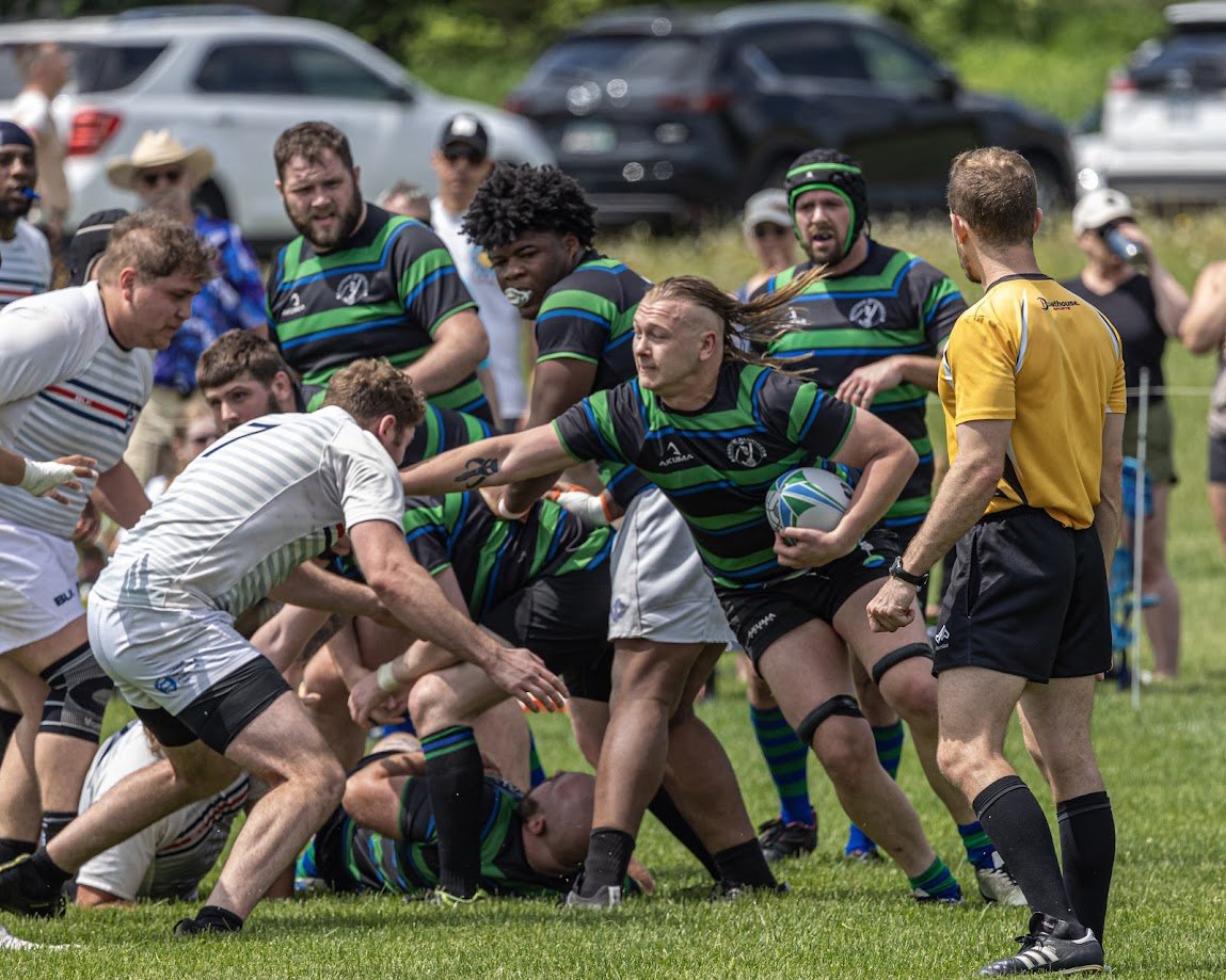 Rugby players engaging in a match on a grassy field, with a referee in a yellow shirt observing, and onlookers in the background near parked cars.