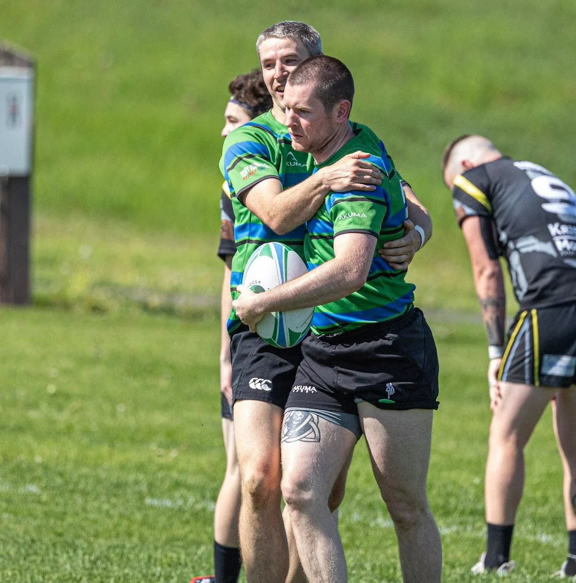 Two rugby players hugging, one holding a rugby ball, on a grassy field during a game.