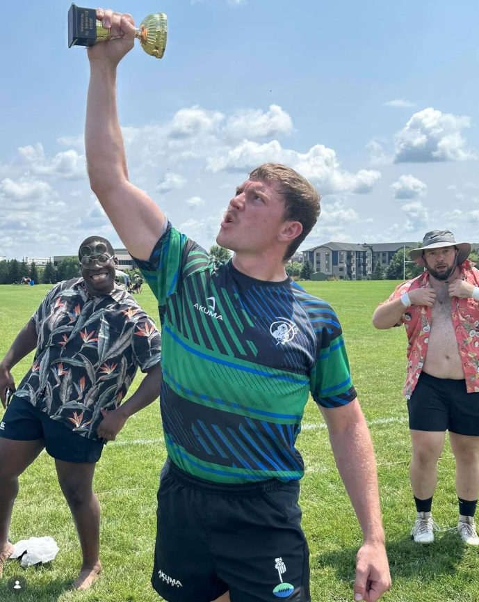 A young man holding a gold trophy up in the air on a grassy field, with two other men in casual summer clothes nearby, under a sunny sky with clouds.