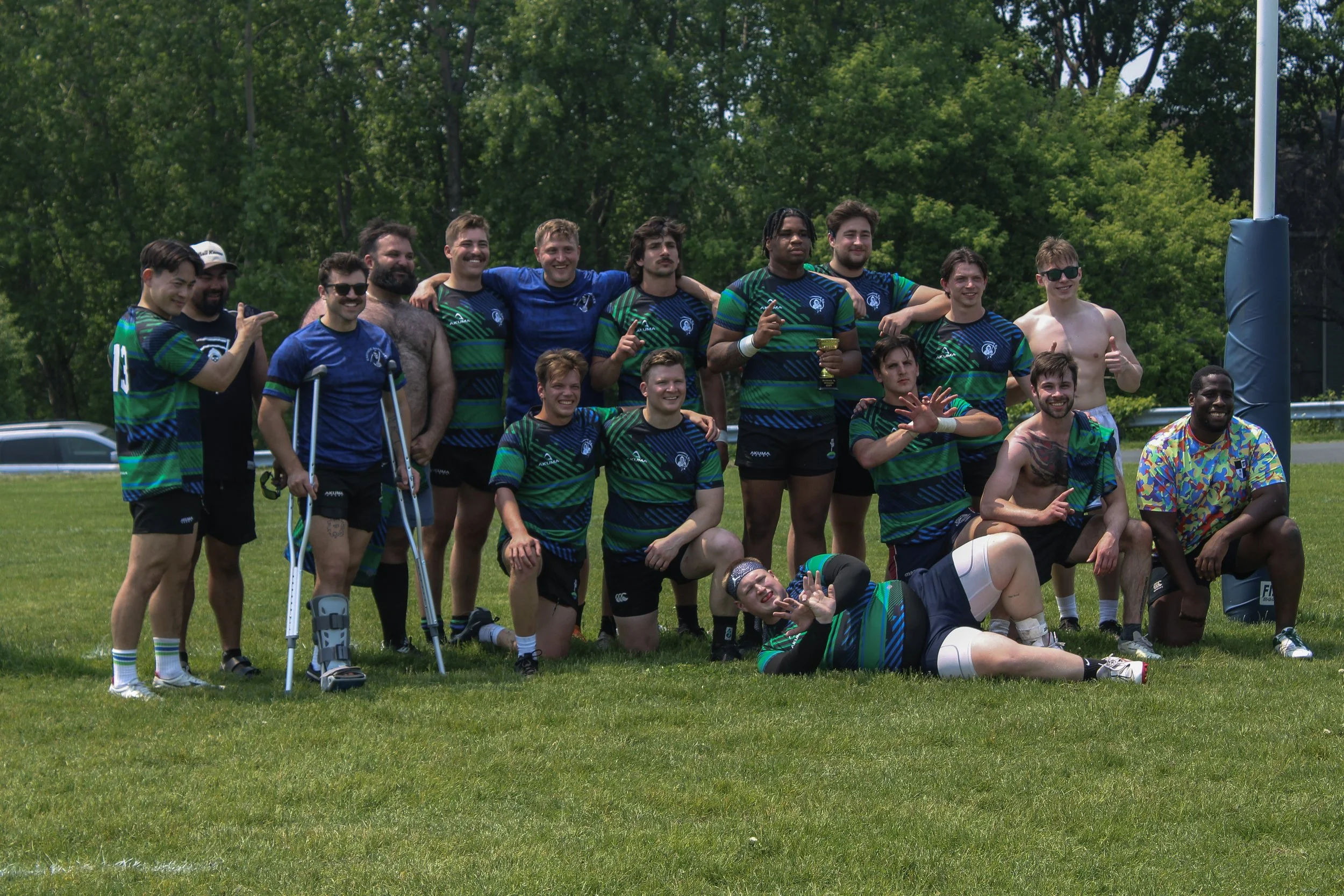 A group of rugby players and supporters pose together after a game on a grassy field, some wearing matching team jerseys and one with crutches, with trees in the background.