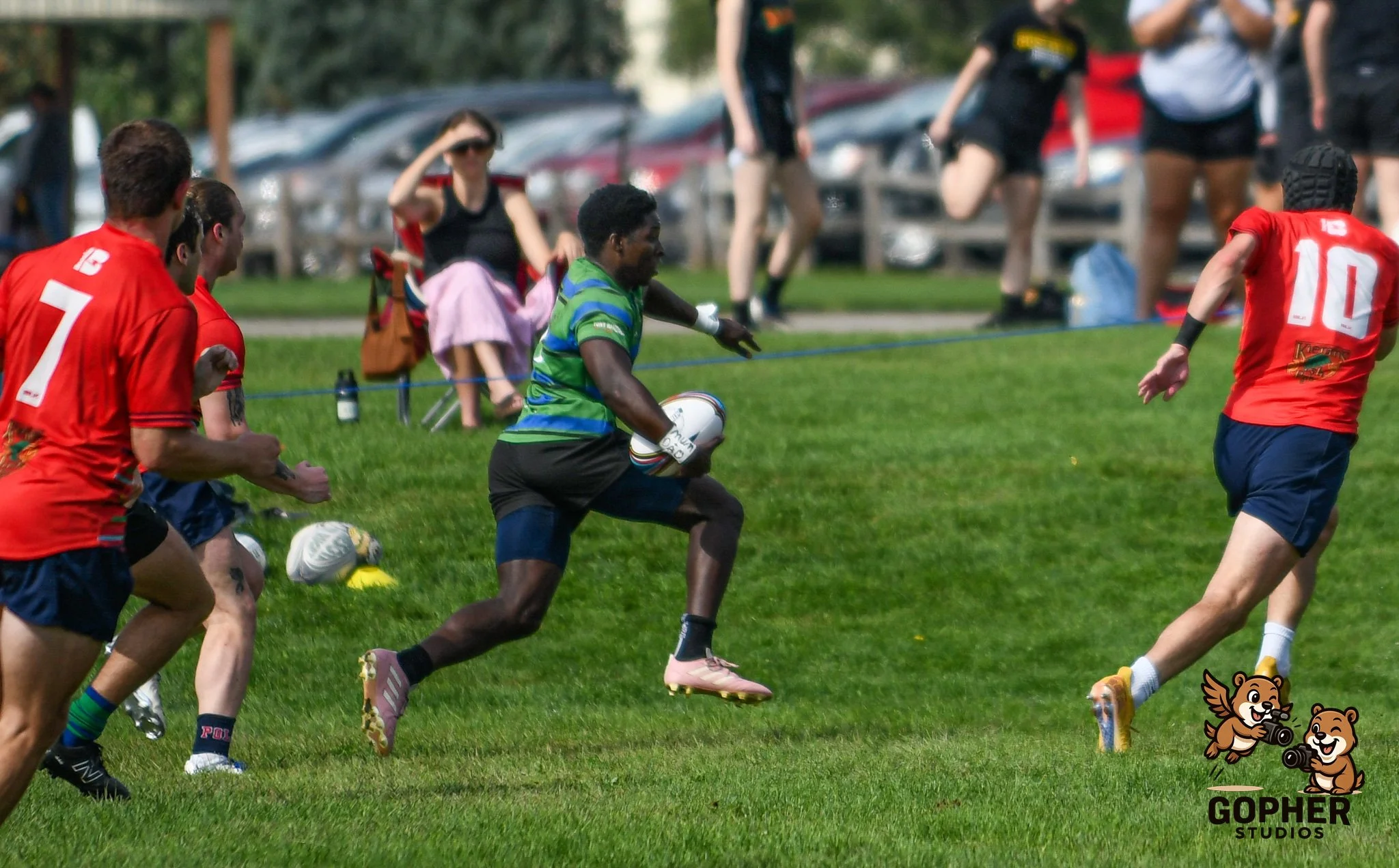 A group of people playing rugby outdoors on a grassy field, with some spectators seated and watching in the background.