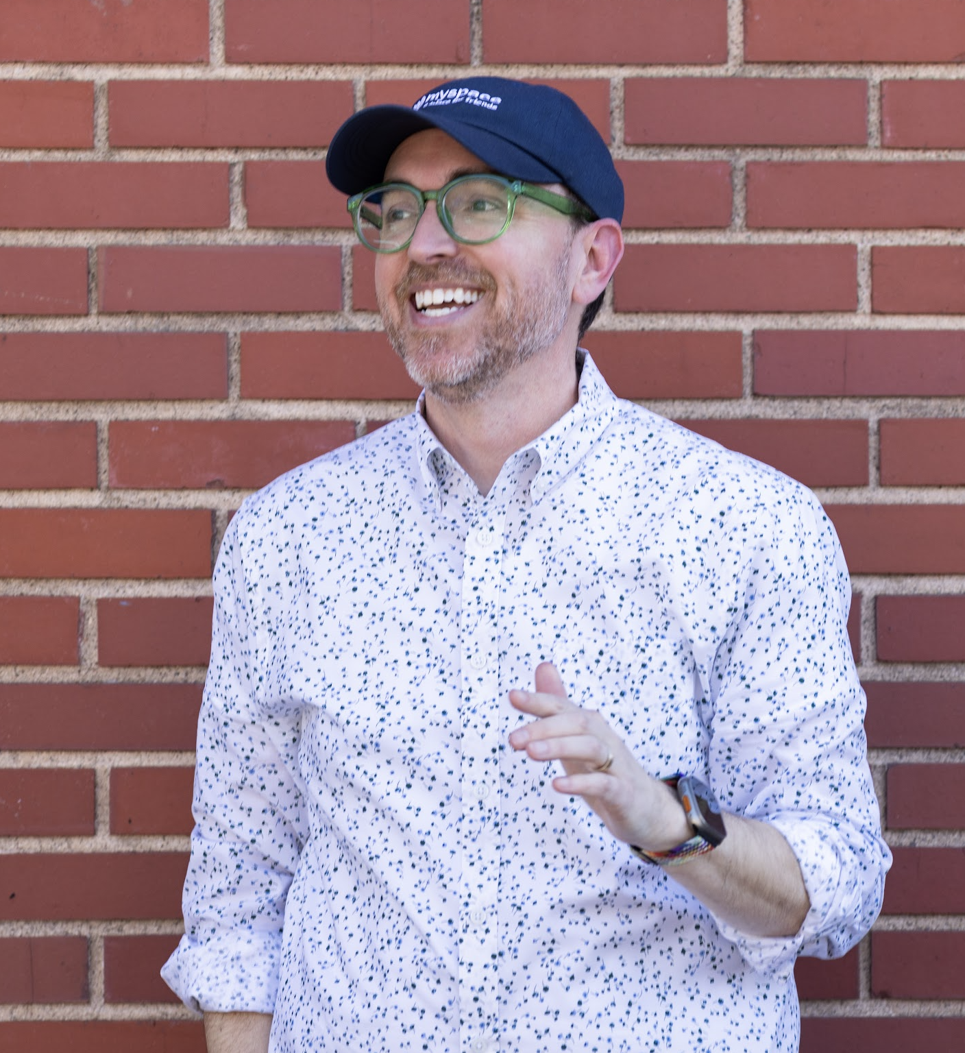 A man with glasses, a beard, and a smile, wearing a dark baseball cap and a white shirt with black speckles, standing in front of a red brick wall.