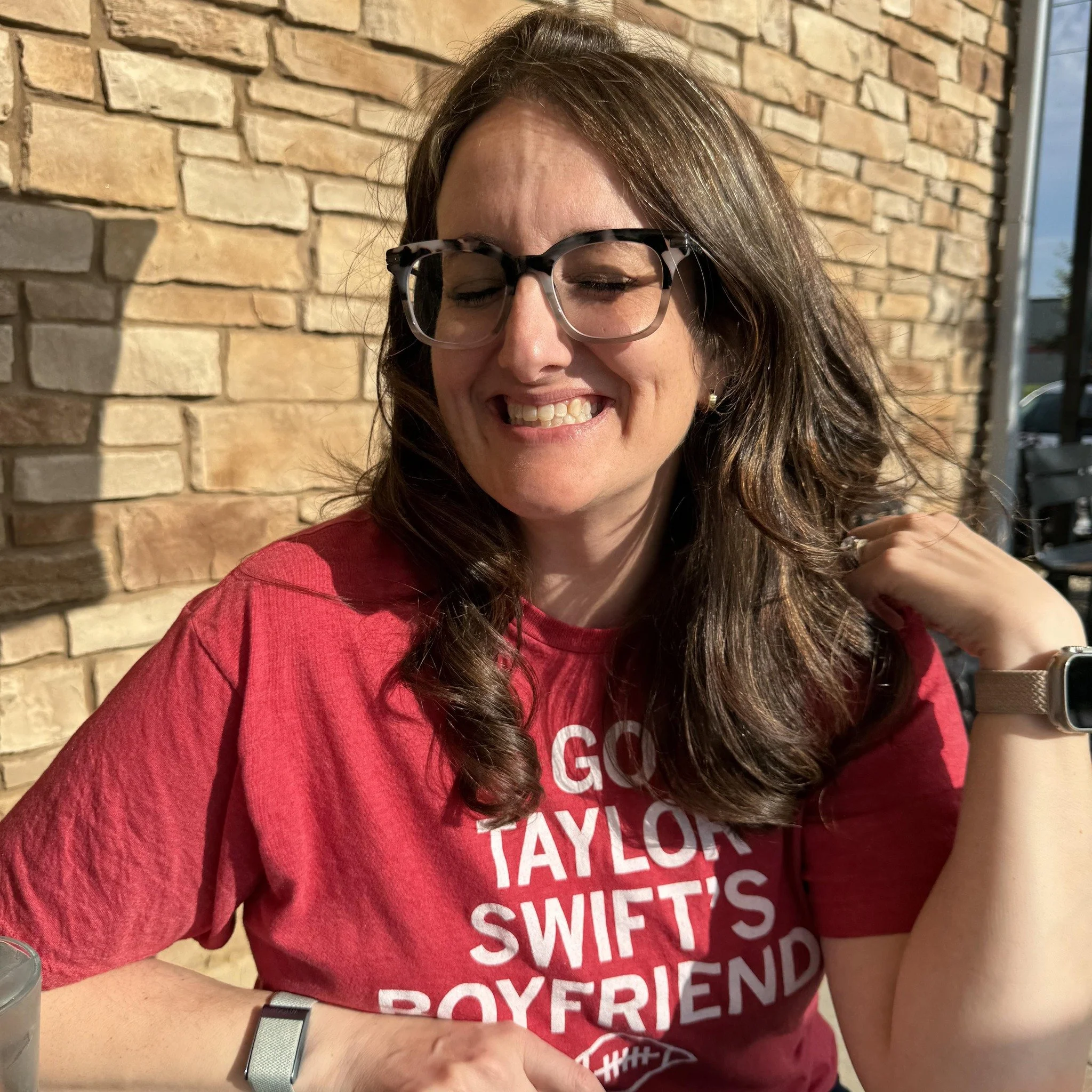 A woman with long brown hair smiling outdoors, wearing glasses and a red shirt with white text, sitting at a table with a brick wall background.