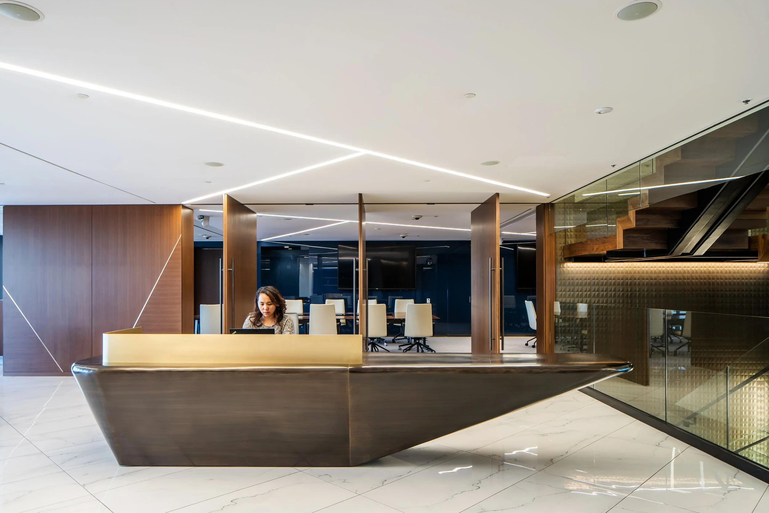 Modern office reception area with a woman at a desk, wooden partitions, white chairs, glass walls, and unique ceiling lighting fixtures.