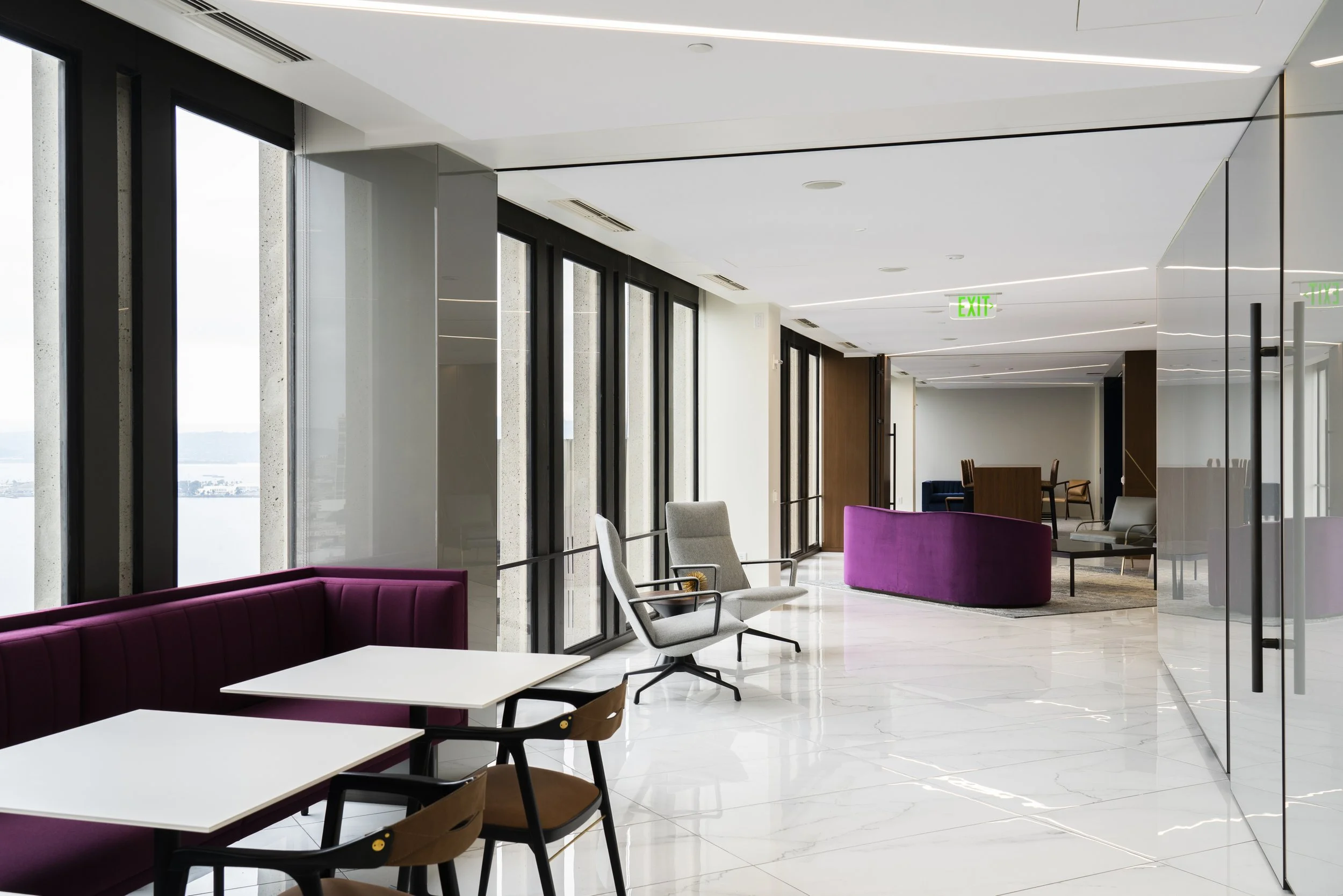 Interior view of a modern building lobby or lounge area with large windows, white walls, and sleek ceiling lighting. There are various chairs and sofas, including purple and grey upholstered seats, and a white table near the window. An exit sign is v