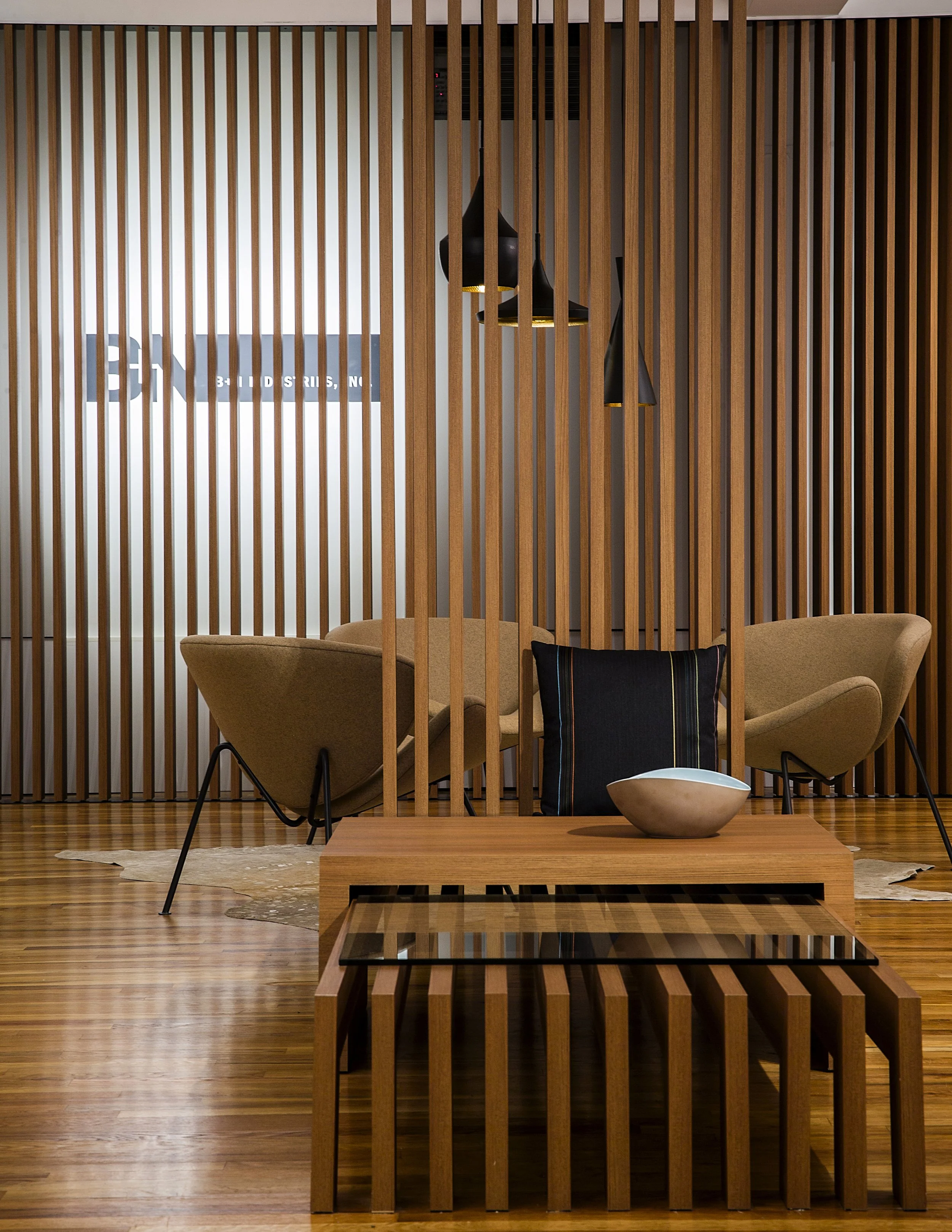 A modern living room featuring a wooden slat partition, a beige sofa with a black pillow, a wooden coffee table with a decorative bowl and a black tray, pendant lights, a sign on the wall, and a cowhide rug on a wooden floor.