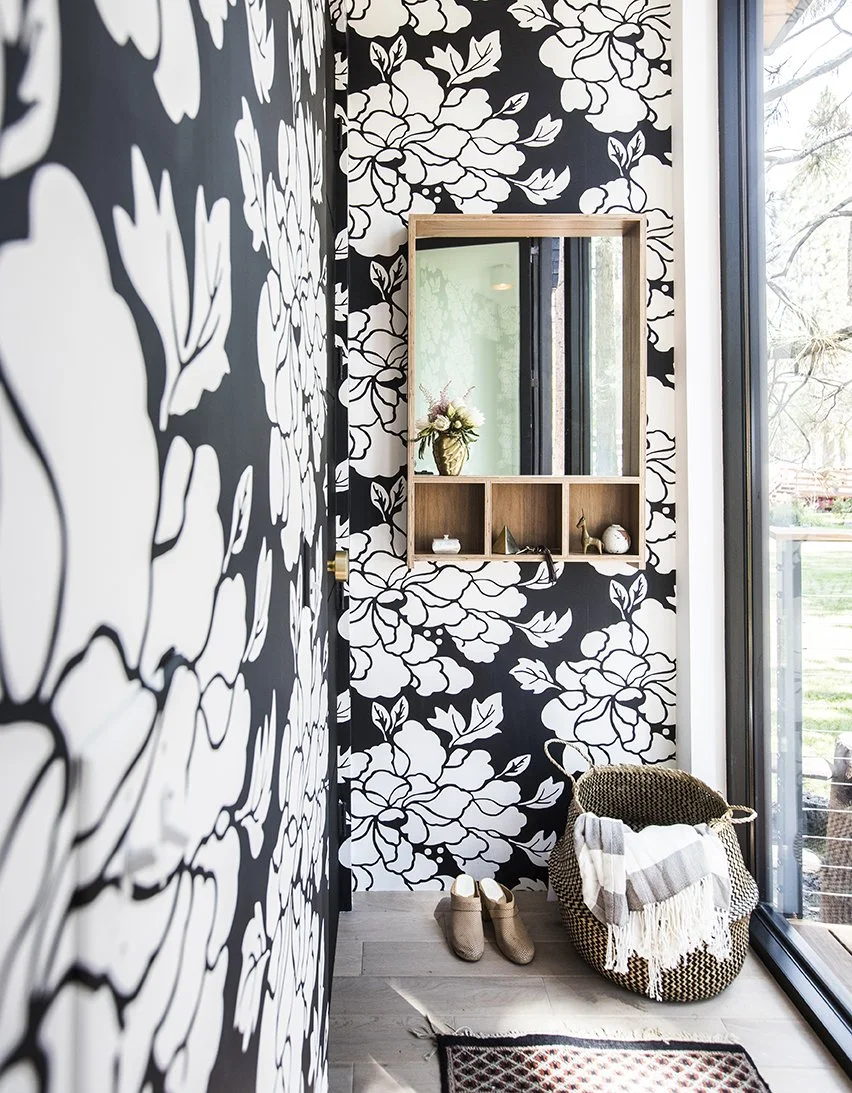 A small entryway with black and white floral wallpaper, a wall mirror with a wooden frame, a wicker basket with a striped blanket, a pair of shoes on the floor, and a large window showing trees outside.
