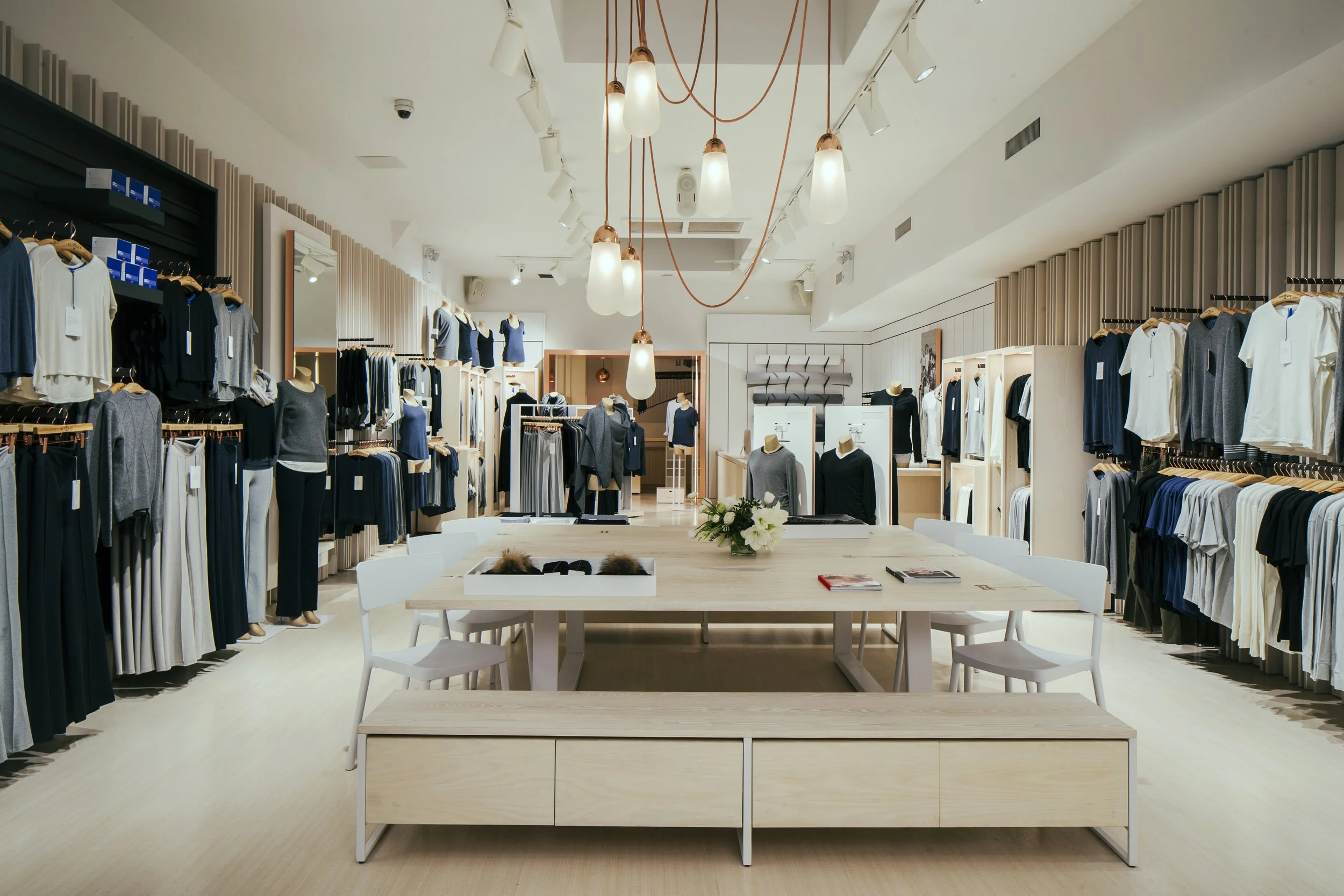 Interior of a clothing store with racks of clothing on both sides, mannequins wearing outfits, a central table with jewelry, and hanging pendant lights.