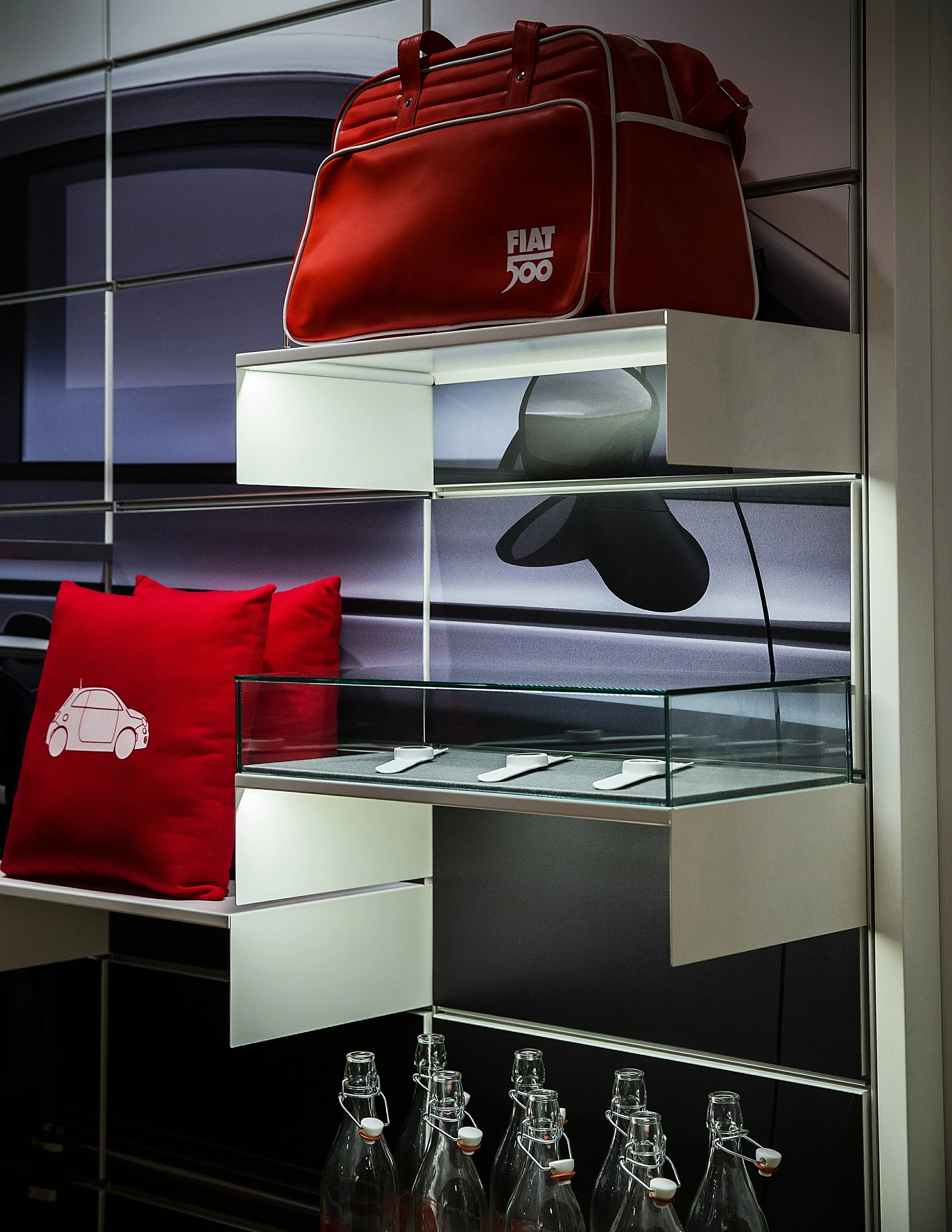 A red Fiat 500 branded duffle bag on a glass and metal display shelf with a white background. Below the shelf are glass bottles with flip-top closures.