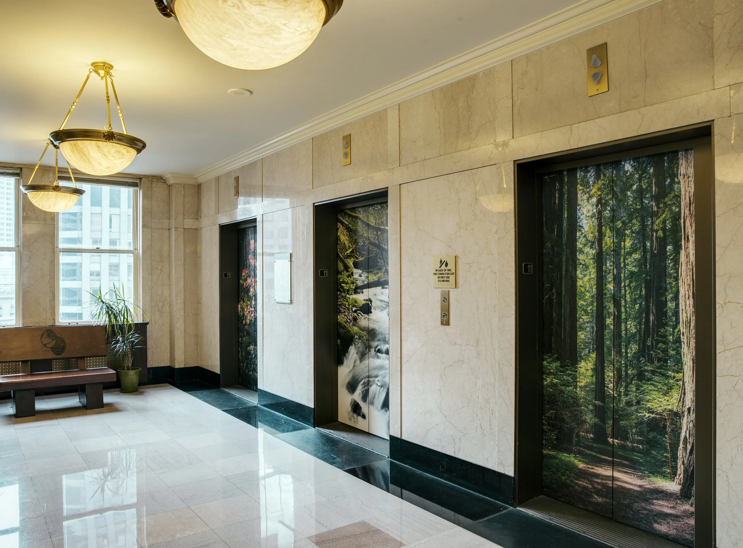 Elevator lobby area with marble walls, two elevator doors with nature-themed artwork, a seating bench, a potted plant, and large windows letting in natural light.