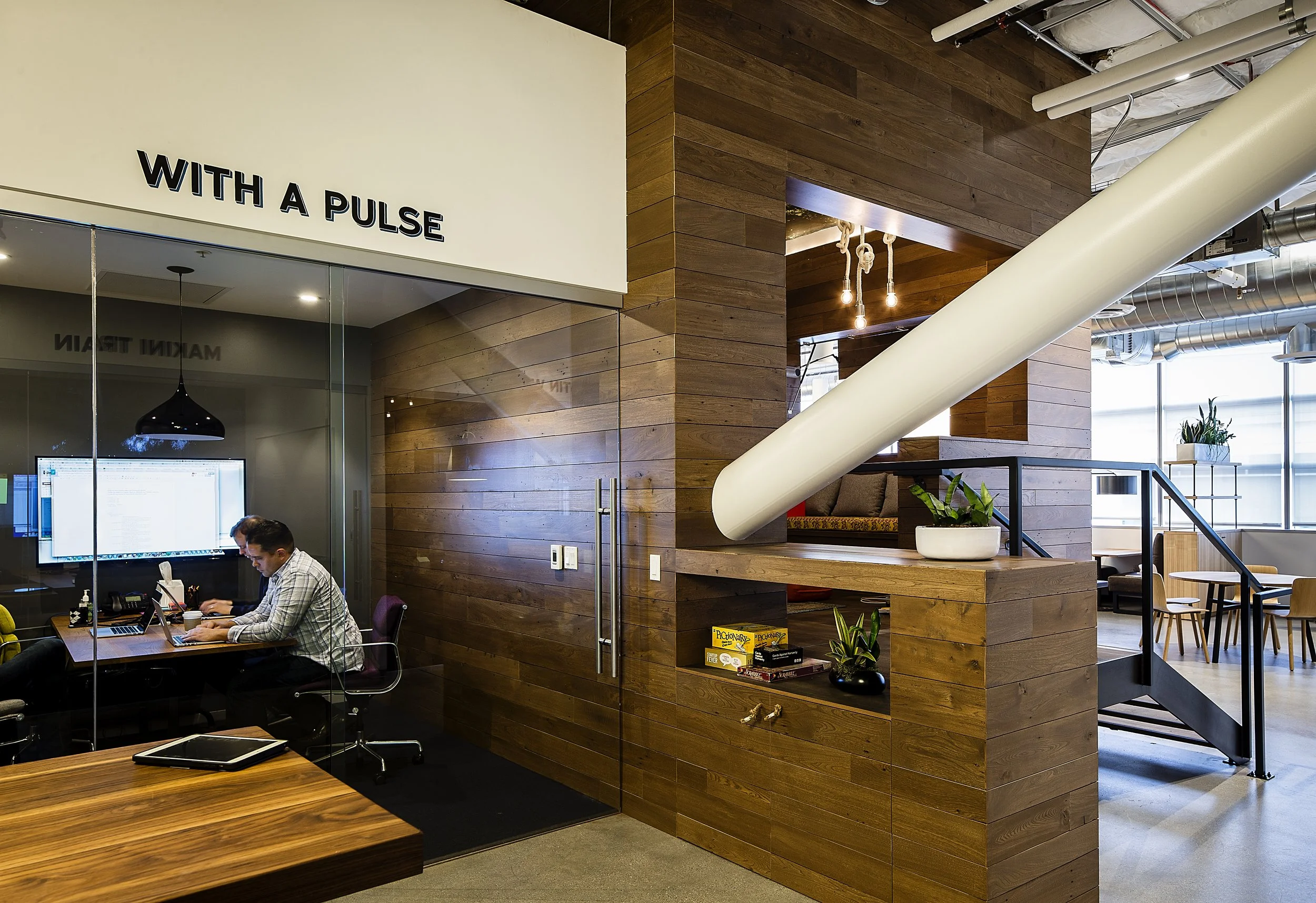 Modern office interior with wood-paneled walls, pendant lighting, and a glass-walled conference room. Two people are working at a desk inside the conference room, and there are potted plants and board games nearby.