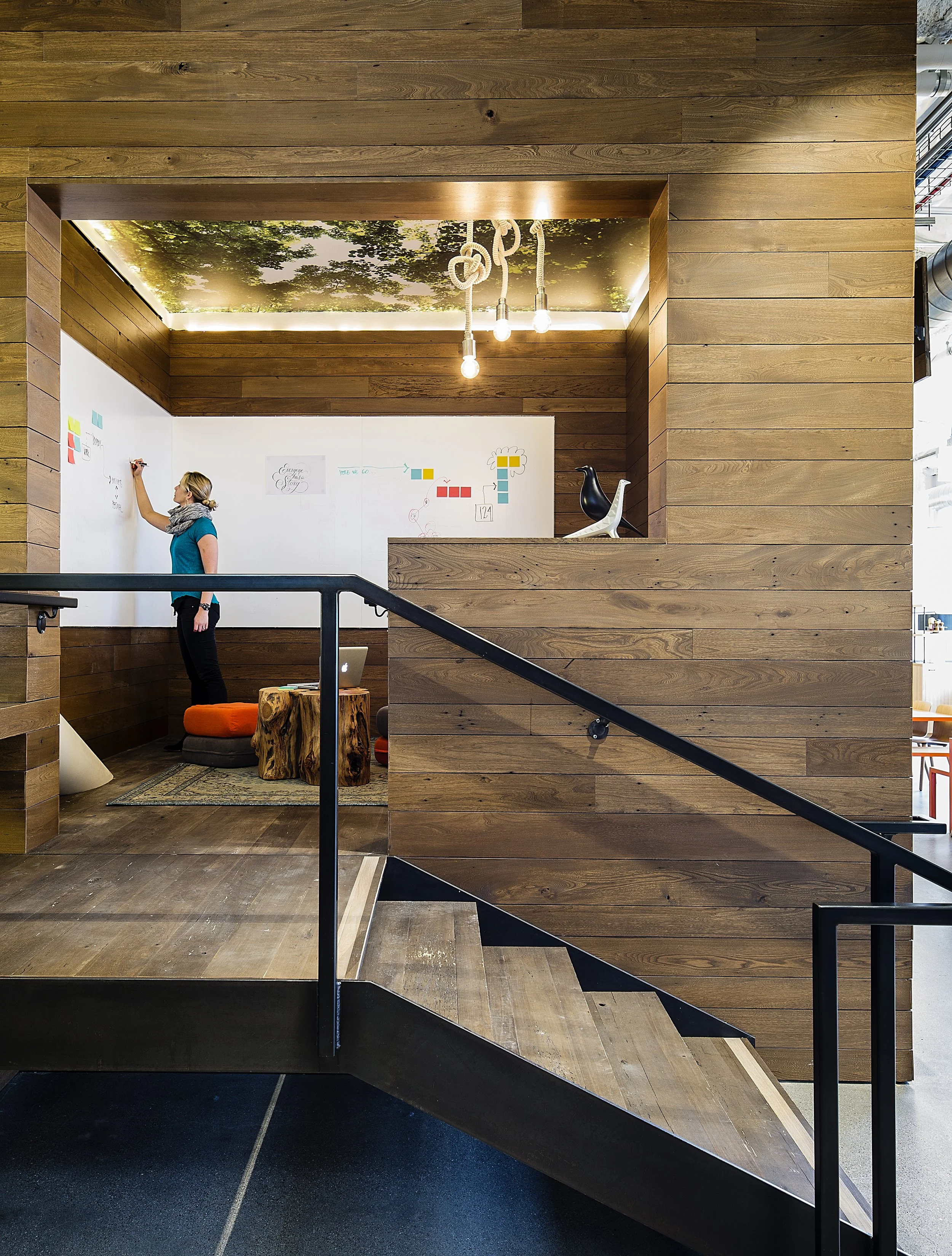 Interior view of a modern workspace with a wooden wall and a whiteboard, featuring a woman writing on the whiteboard, and decorative bird sculptures on the ledge. A ceiling with tree leaf pattern and hanging light bulbs is visible in the background.