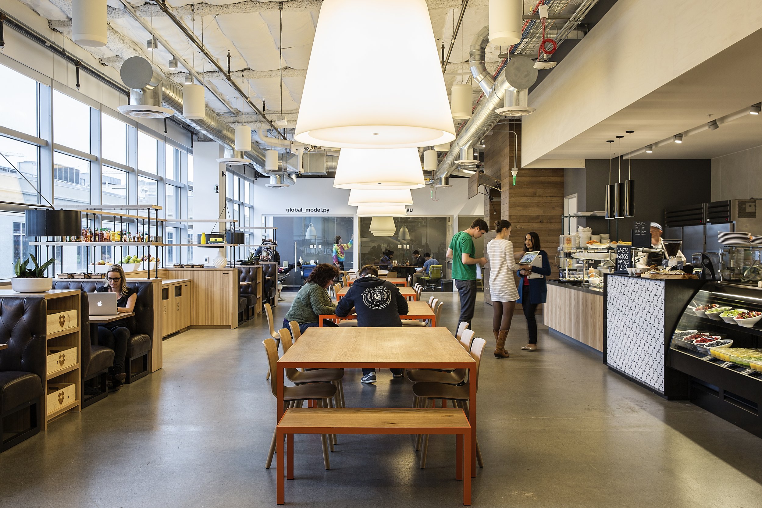 Interior of a modern cafe with large arched windows, wood accents, and contemporary lighting. Customers are seated at tables, some working on laptops, while staff take orders at the counter.