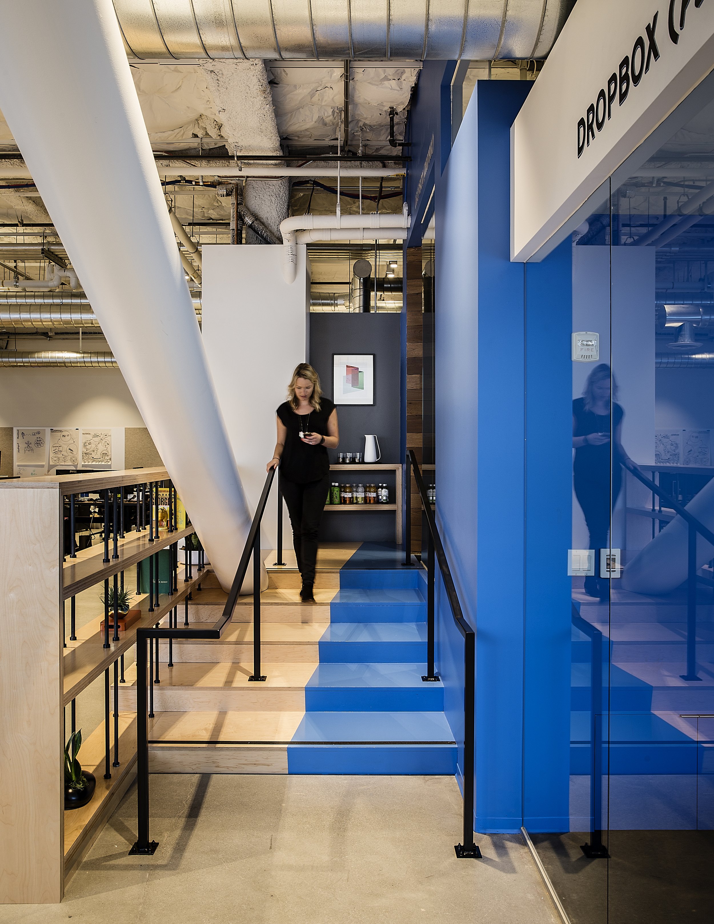 Interior of an office space featuring a staircase with a black metal railing, a blue partition wall labeled 'DROP BOX,' an illuminated sign, and a woman with blond hair wearing a black dress walking down the stairs. The background includes exposed du