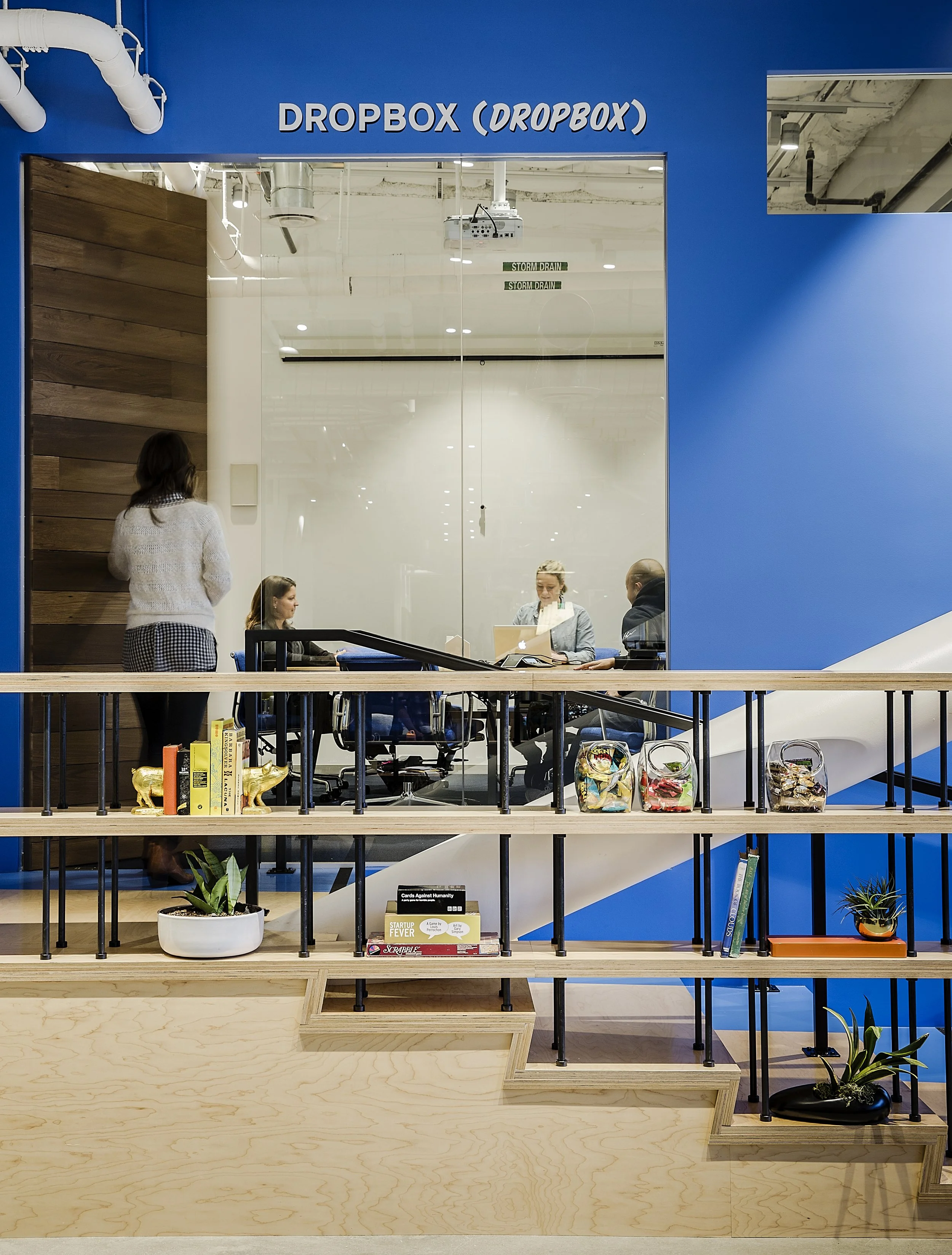 View of a modern office space with a glass-walled meeting room. The room has a sign reading 'DROPBOX (DROBOX)' and shows people sitting and standing inside. The foreground has a light-colored wood counter with books, plants, and glass jars filled wit