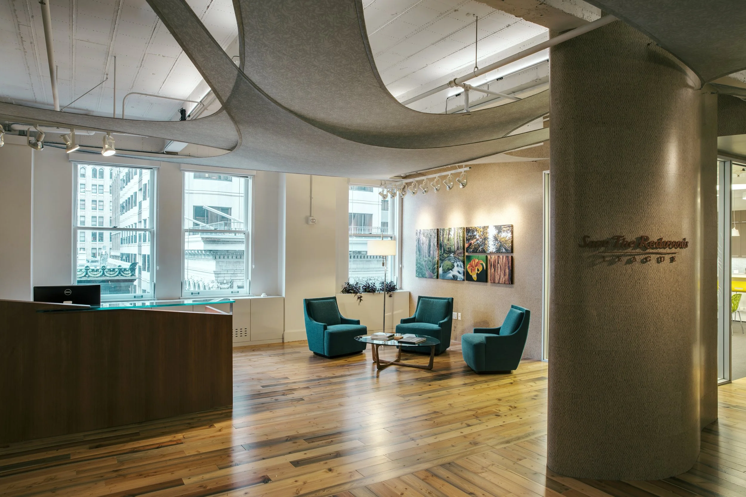 Modern office or lounge area with teal armchairs, wooden flooring, large windows, and wall artwork.