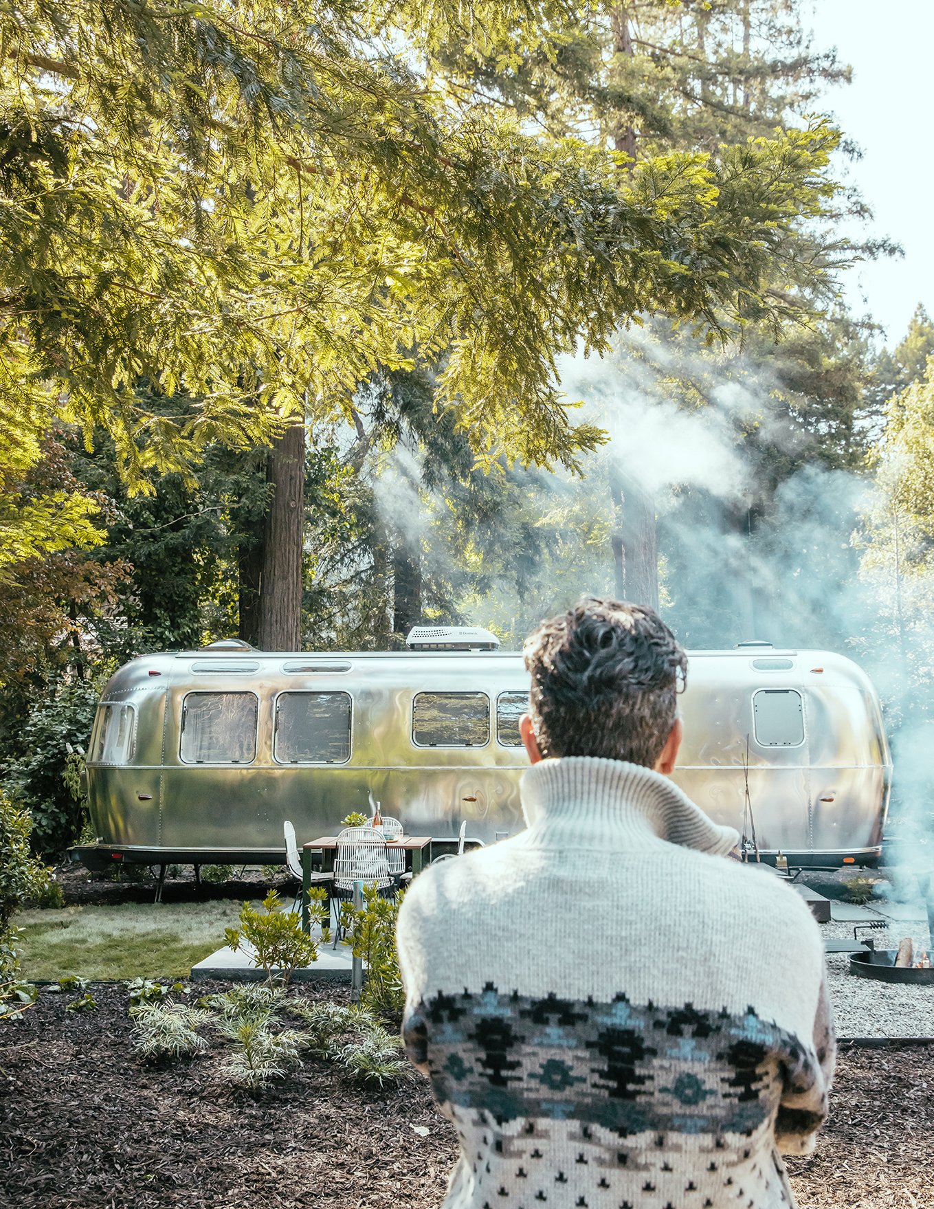A person with dark hair and a patterned sweater is standing outdoors in front of a shiny, vintage Airstream trailer in a wooded area with tall trees and sunlight filtering through. Smoke or steam is rising behind the trailer.