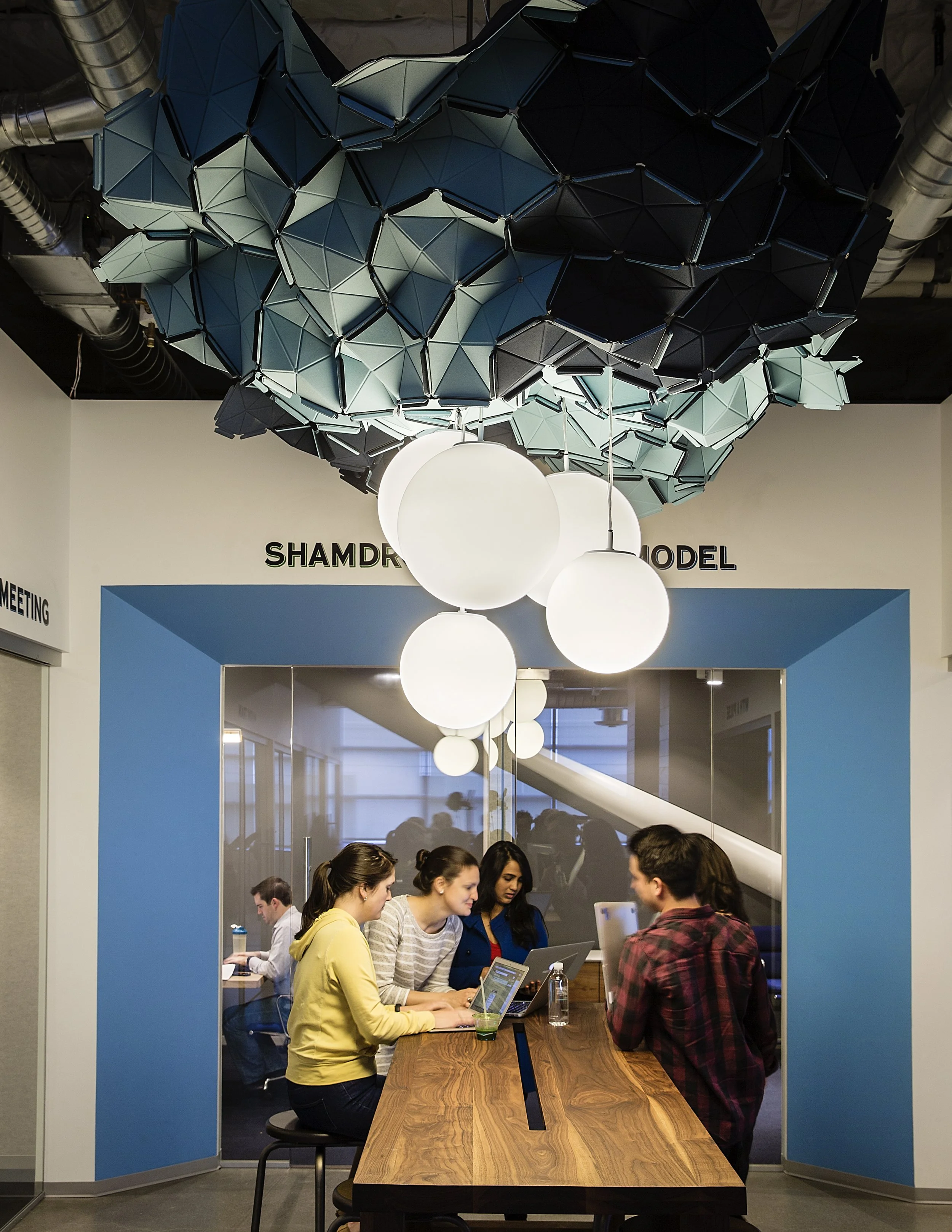 A group of people gathered around a computer at a long wooden table inside a modern office or coworking space. Behind them, there are three large white spherical pendant lights hanging from the ceiling, and a blue wall with signs reading 'SHAMDR' and