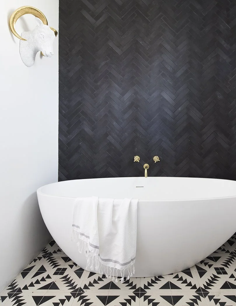 A modern bathroom with a white freestanding bathtub, a black chevron tile wall, and black and white geometric patterned floor tiles. A white towel hangs over the tub, and a wall-mounted white animal head with gold horns decorates the white wall.