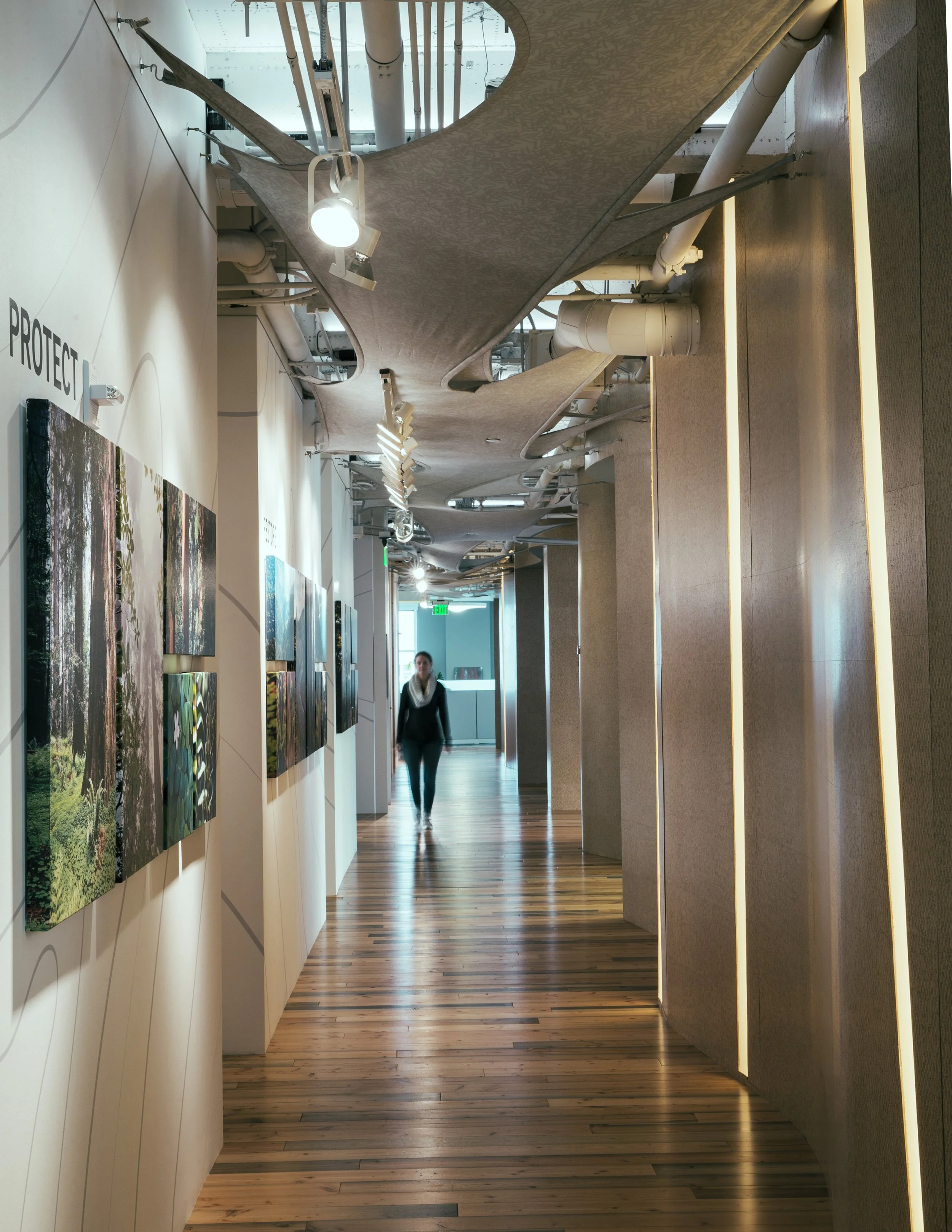 A modern art gallery with a wooden floor, white walls on the left displaying colorful landscape photographs, and beige textured panels on the right. Overhead track lighting illuminates the space, and a woman walks inside the gallery.