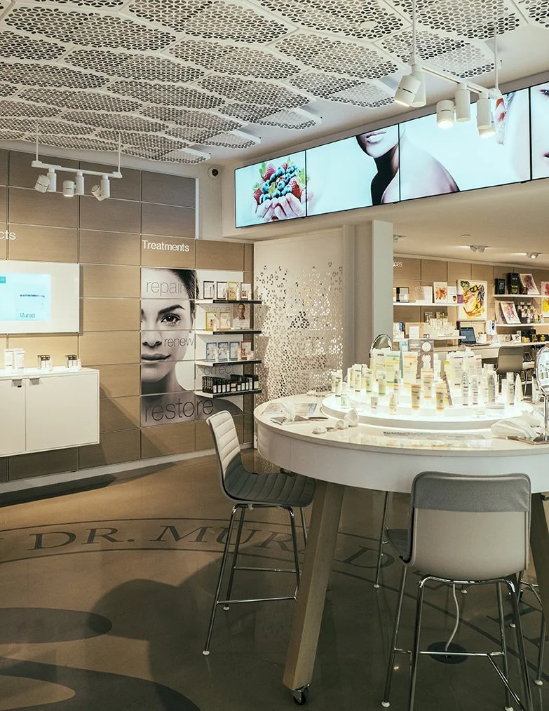 Interior of a cosmetics store with skincare products on display, digital screens showing promotional images, and seating around a circular display table.