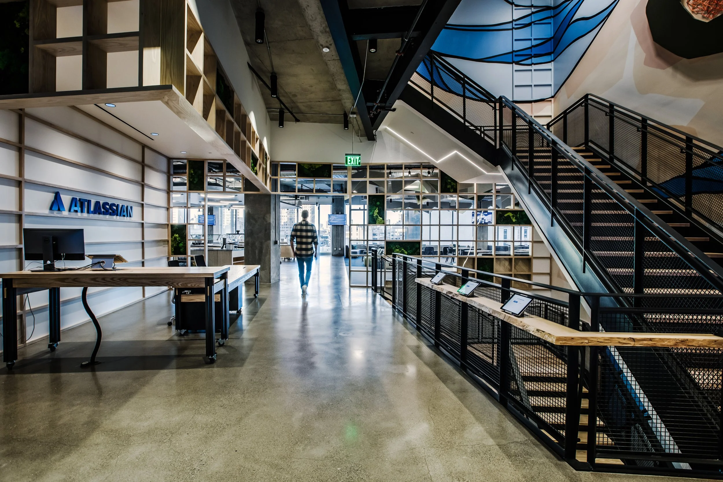 Interior of an office building with a modern design, featuring wooden shelving, a staircase with black railings, large windows, and a person walking towards the exit.