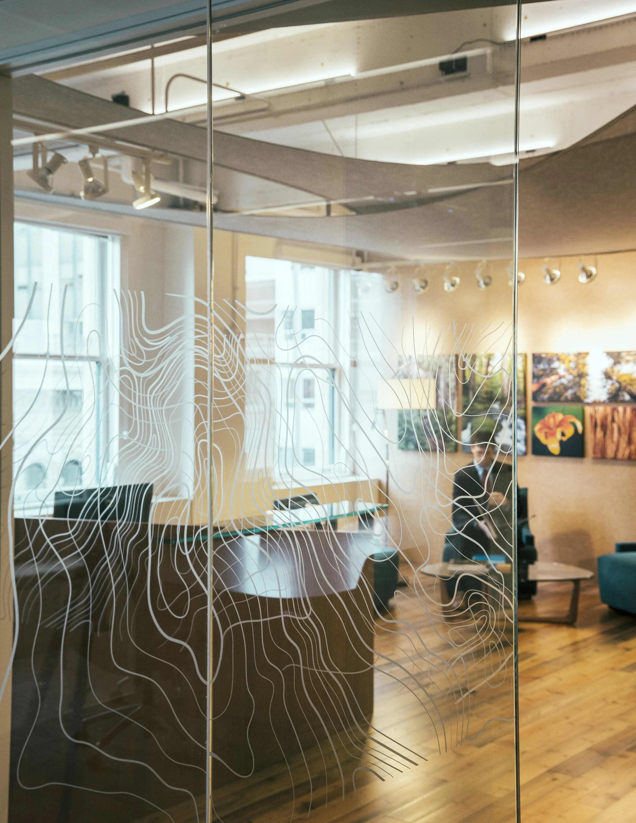 Reflection of an art gallery or studio in a glass door with abstract white line art design, showing artwork on walls and someone sitting at a desk inside.