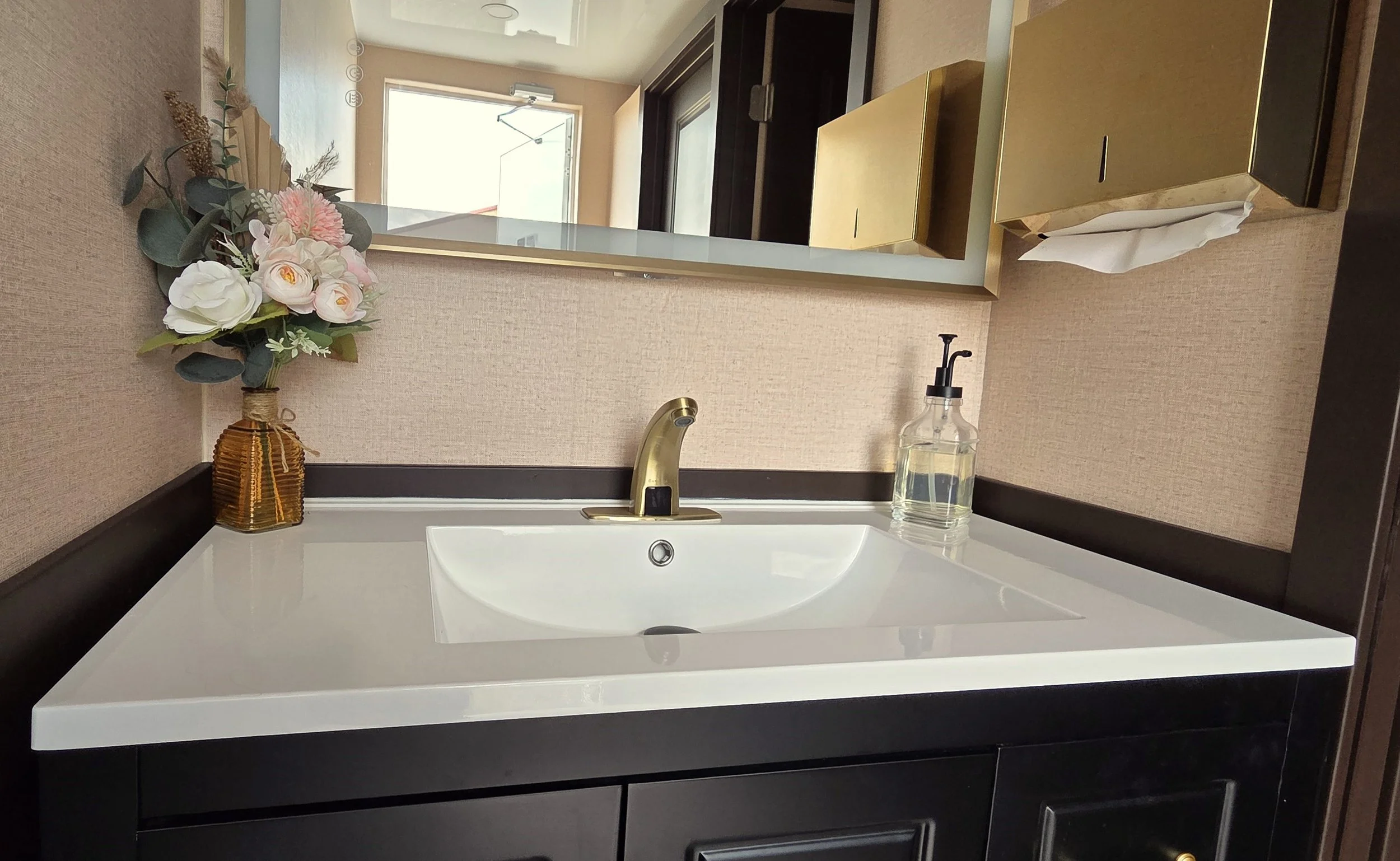 A FIESTA LUXURY RENTAL bathroom mirror with a beige wall behind it, reflecting a door and window. On the bathroom countertop, there is a flower arrangement in a brown vase, a soap dispenser, and a gold box.