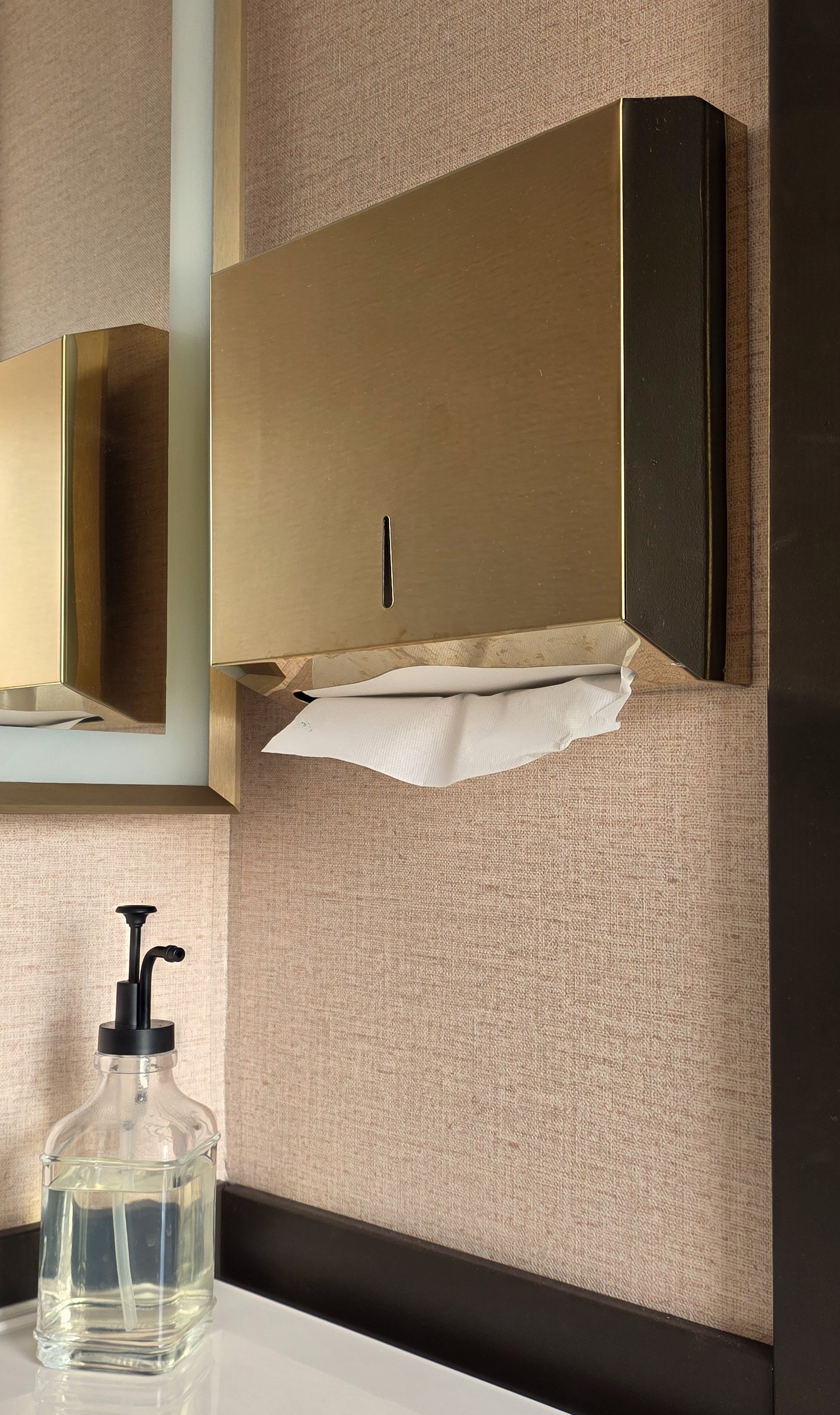A beige wall with a brass tissue dispenser, a mirror, a Gold  sink faucet, and a bottle of hand soap on a white countertop.