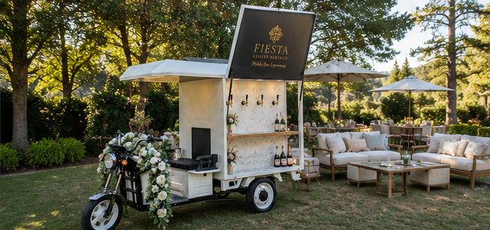 A FIESTA LUXURY mobile bar cart attached to a motorcycle, decorated with white flowers, at an outdoor event with seating and umbrellas in a park setting during sunset.