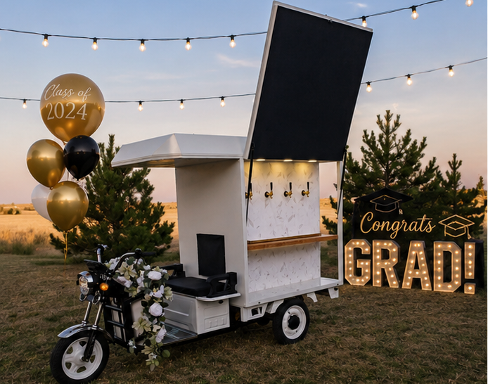 Outdoor graduation celebration with a decorated fiesta luxury rentals electric beverage cart, gold and black balloons reading 'Class of 2026,' a 'Congrats Grad' sign, and string lights at sunset.