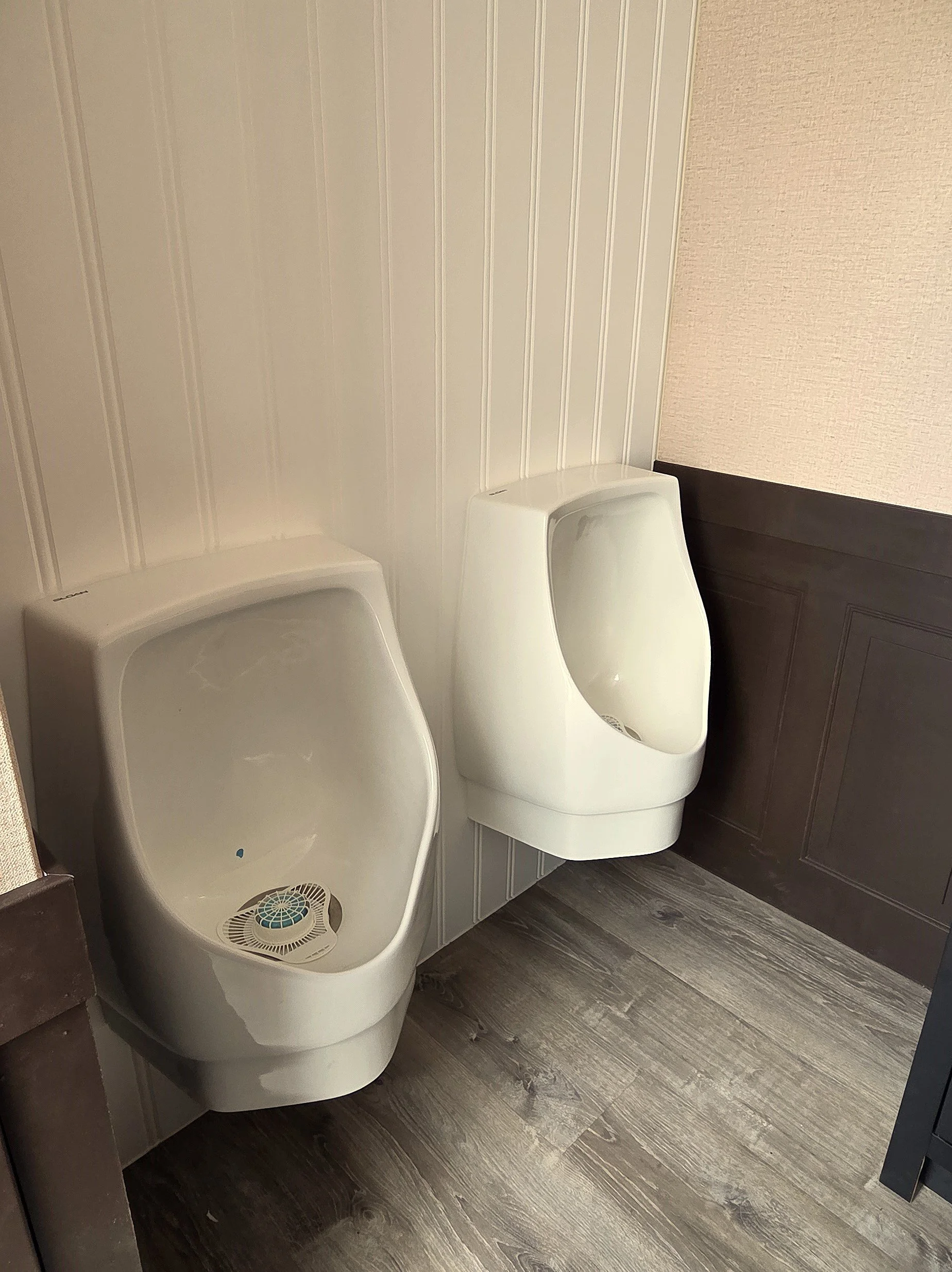 Two white urinals with a low partition between them in a bathroom with brown wood-look floor and textured light beige wall.