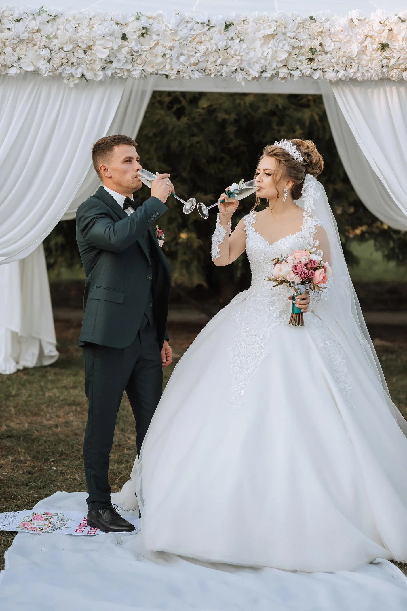 A bride and groom in wedding attire standing on a white fabric platform beneath a floral arch, drinking champagne. The bride wears a white lace wedding gown, a tiara, and holds a bouquet of pink roses. The groom wears a dark green suit with a bowtie.