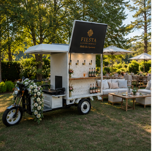 A mobile bar setup with a black sign that reads "Fiesta" on a sunny outdoor lawn, decorated with flowers and surrounded by outdoor furniture and umbrellas.
