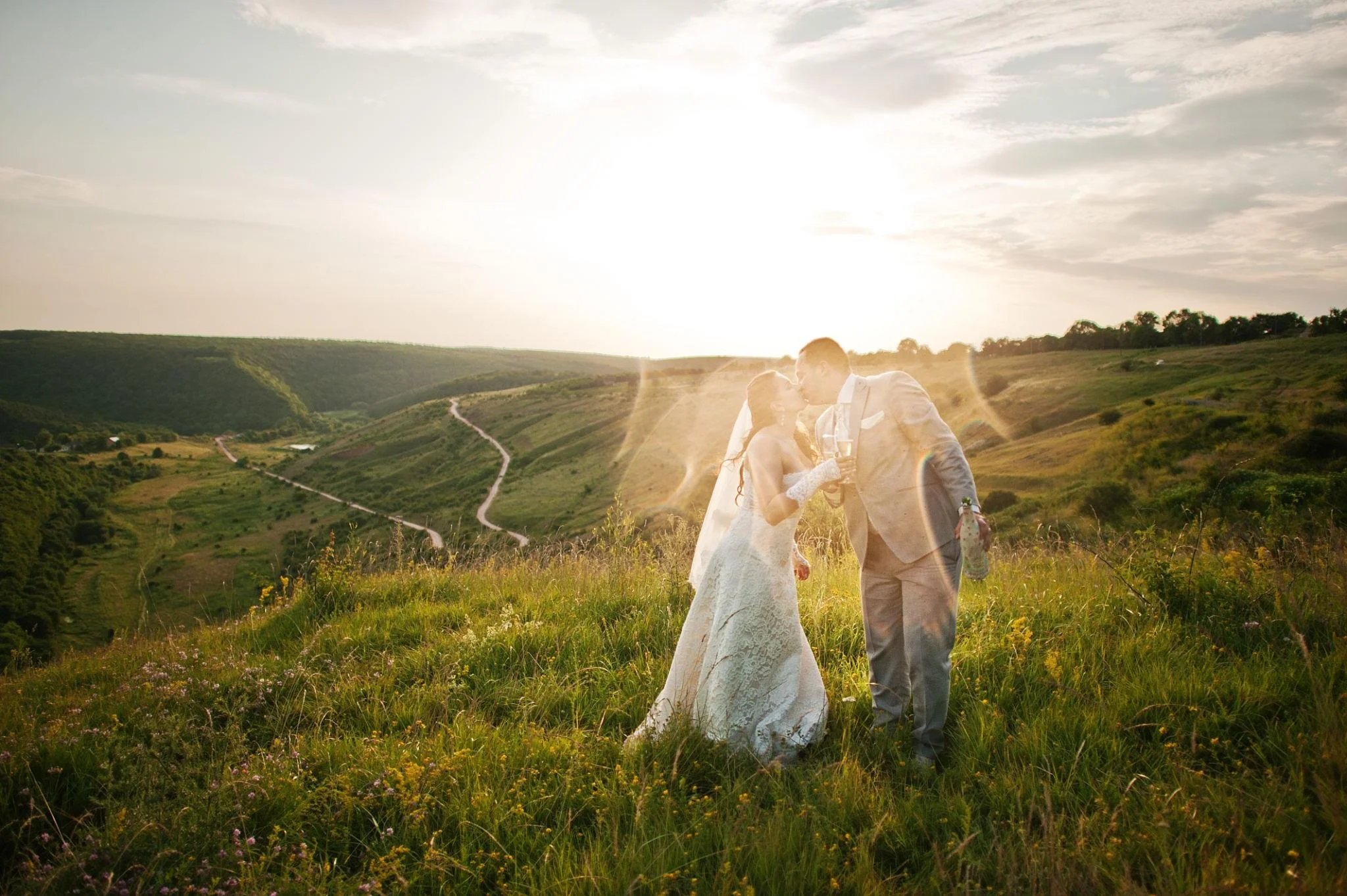 A couple dressed in wedding attire sharing a kiss in a lush green field with scenic hills and a winding dirt path, during sunset with warm sunlight celebrating with fiesta luxury rentals