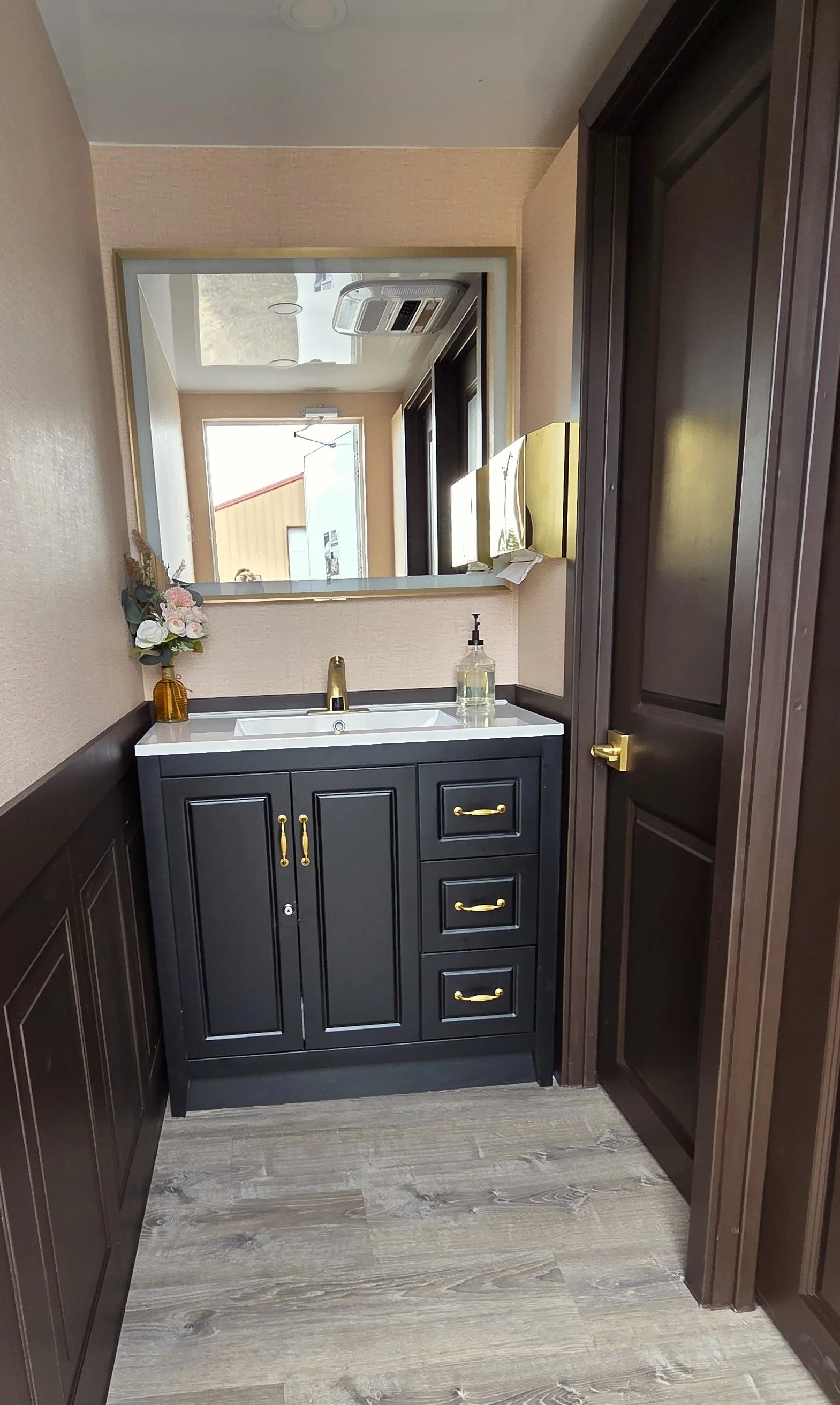 Small bathroom vanity with black cabinets, gold hardware, a white sink, a gold faucet, and decorative flowers in a vase. A mirror is above the vanity, reflecting a door, window, and some objects.